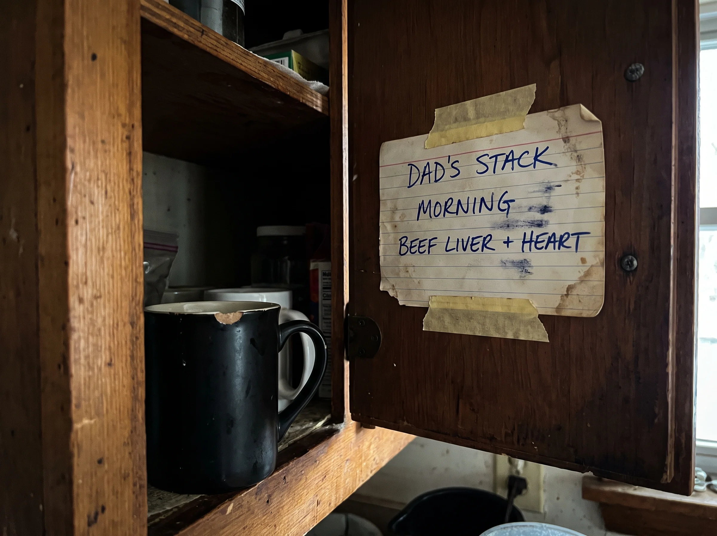 A weathered index card taped inside a kitchen cabinet door, three handwritten lines in blue ballpoint pen, slightly smudged. 