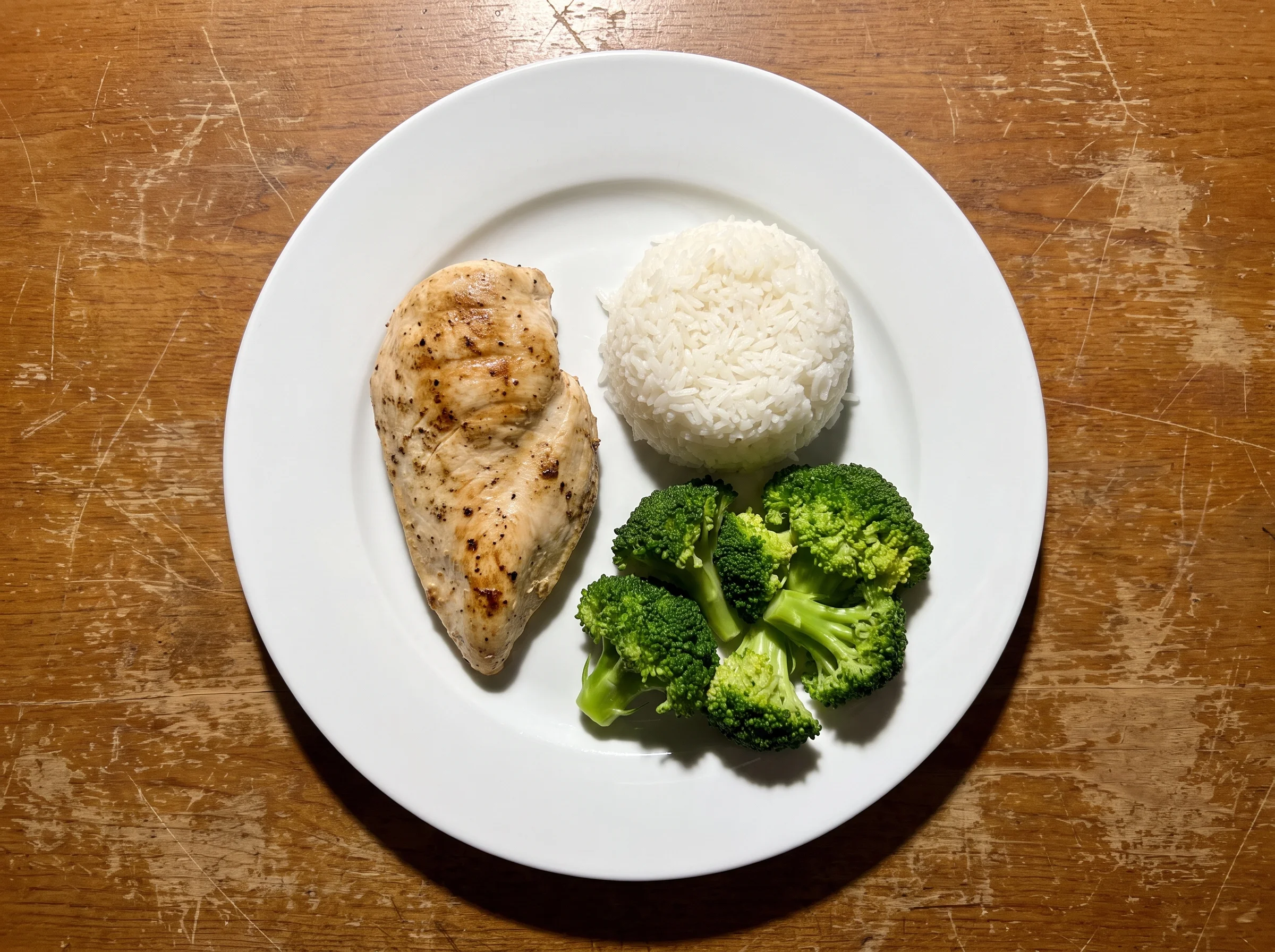 A dinner plate with grilled chicken breast, plain white rice, and steamed broccoli, shot from directly above on a wooden tabl