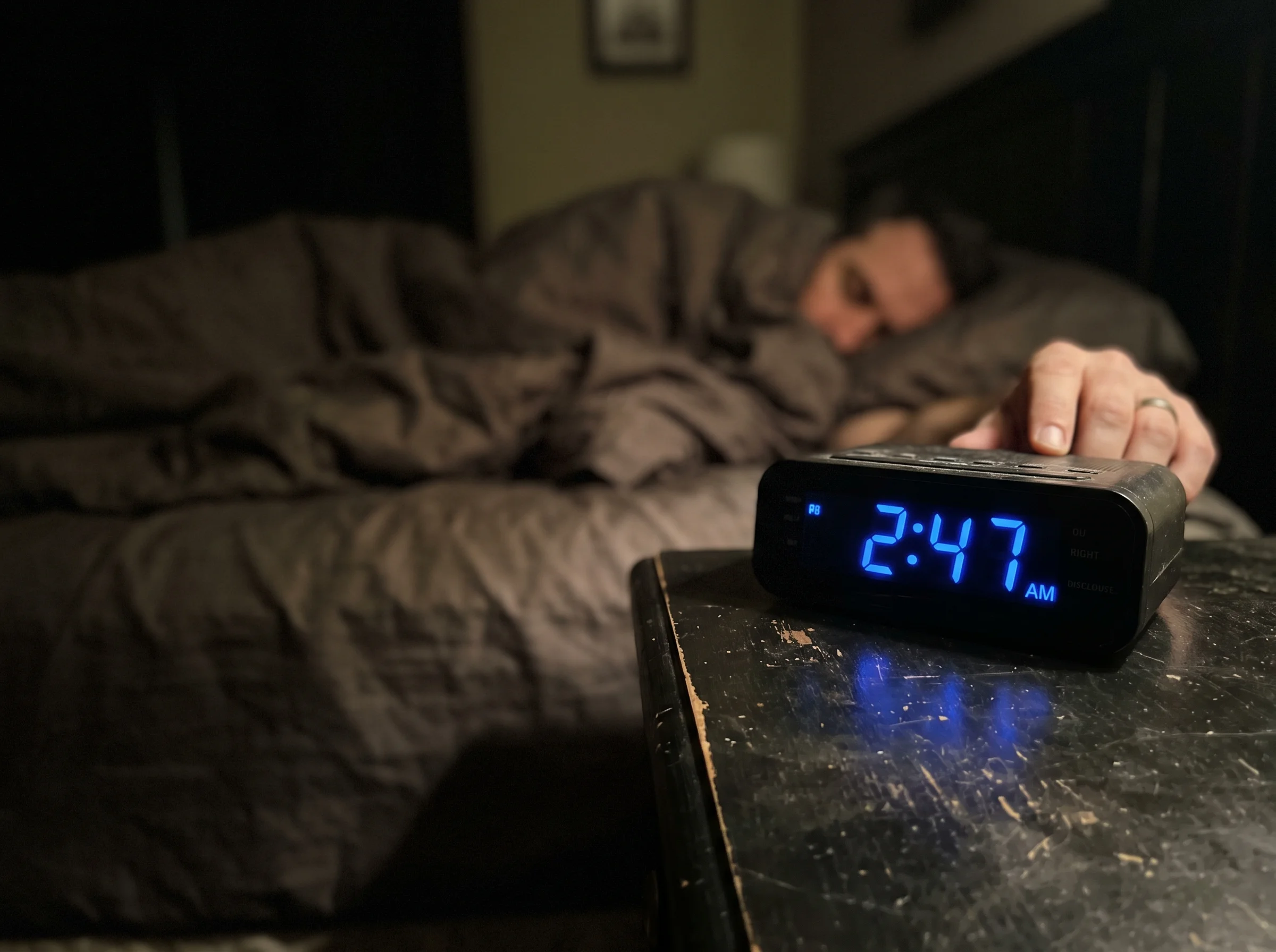 A close-up of a man's alarm clock at 2:47am on the nightstand, blue digital numbers glowing in a dark bedroom, blurred outlin