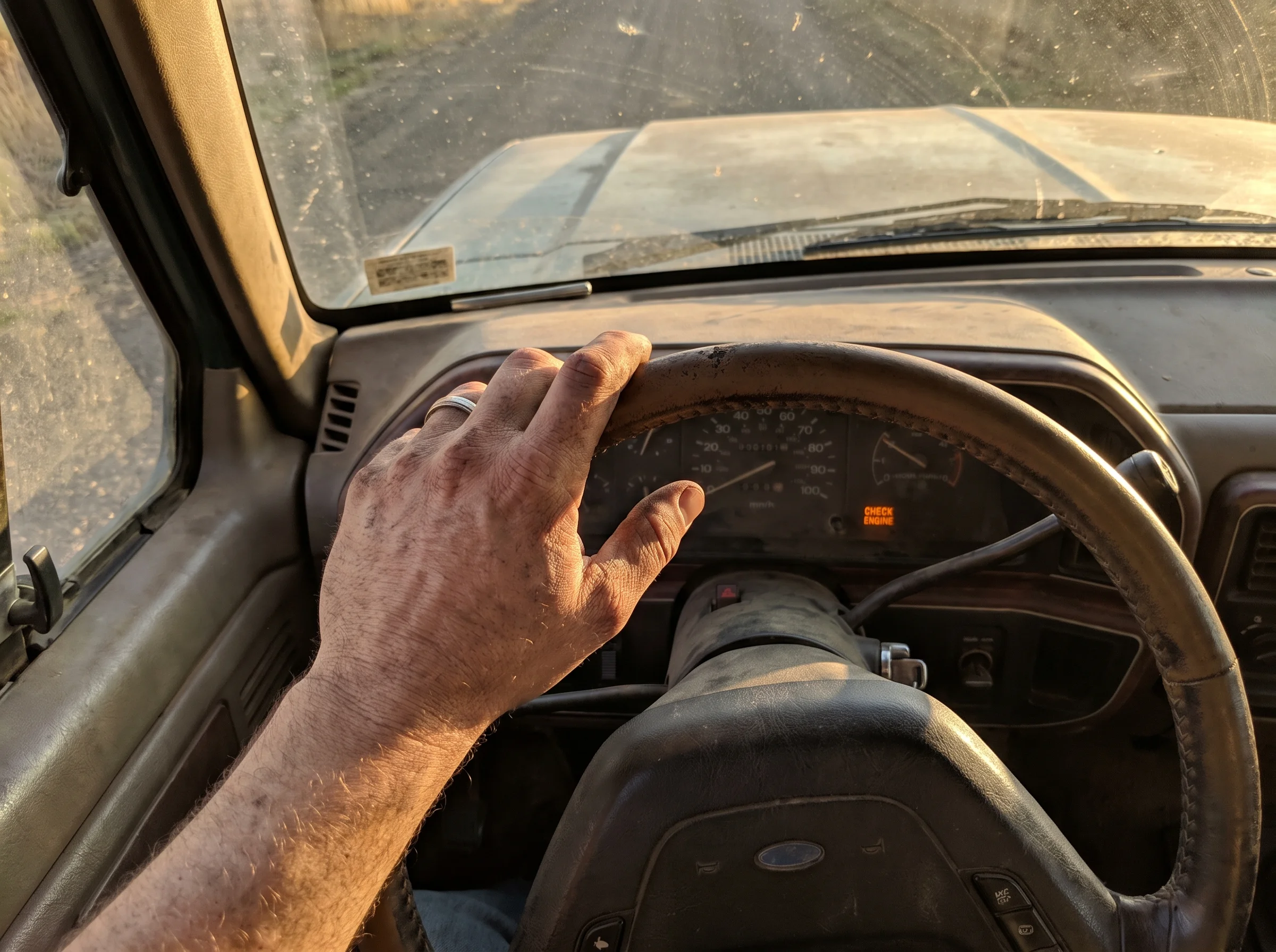 Close-up of a rugged man's hand on a truck steering wheel at 3pm, late afternoon sun cutting through the windshield, dashboar