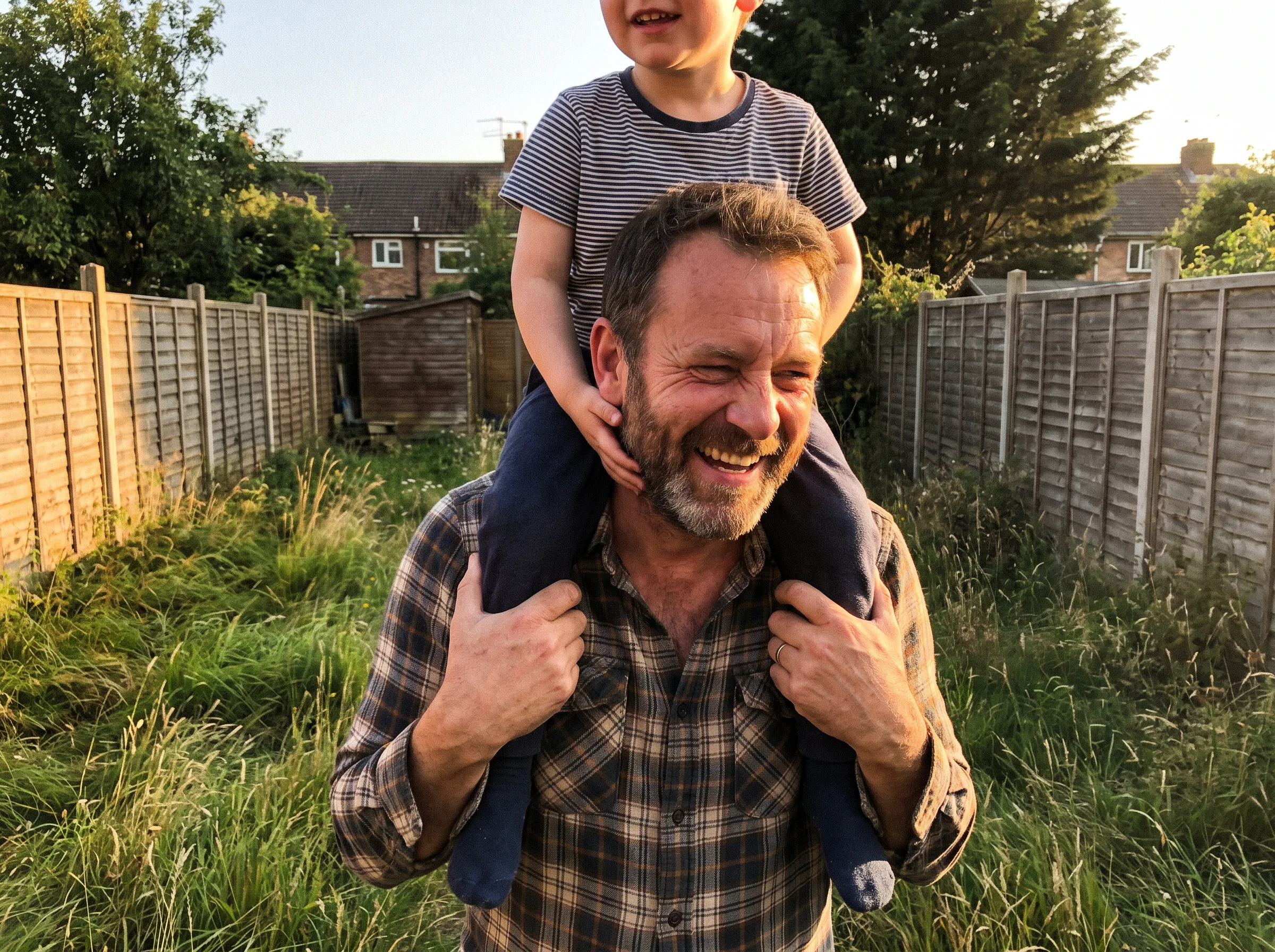 A man in his early 50s carrying a young boy on his shoulders through a backyard at golden hour, both laughing, real weight in