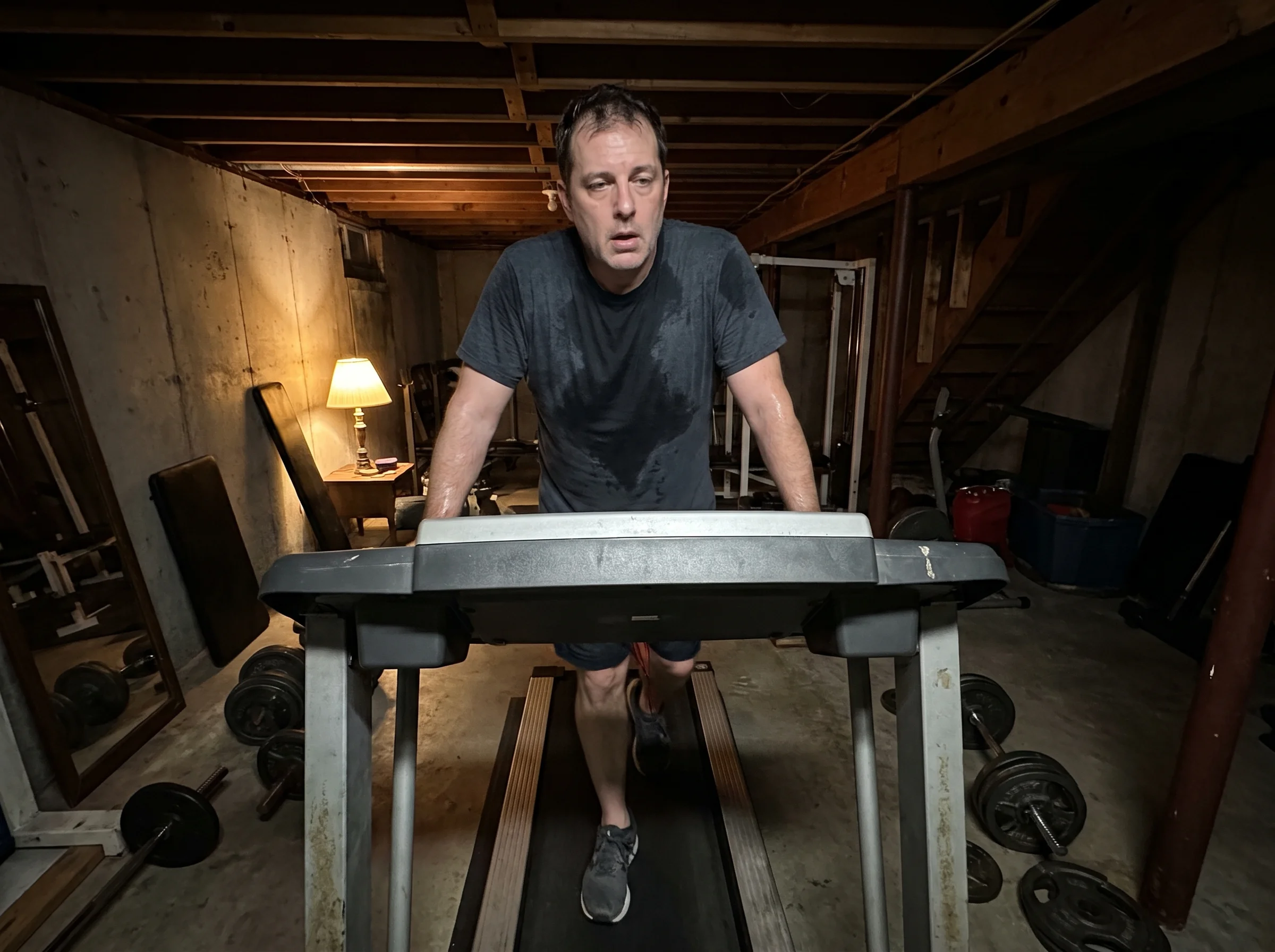 A man in his late 40s on a treadmill in a dim home gym at 6am, face slack with fatigue, sweat on his shirt, staring at nothin