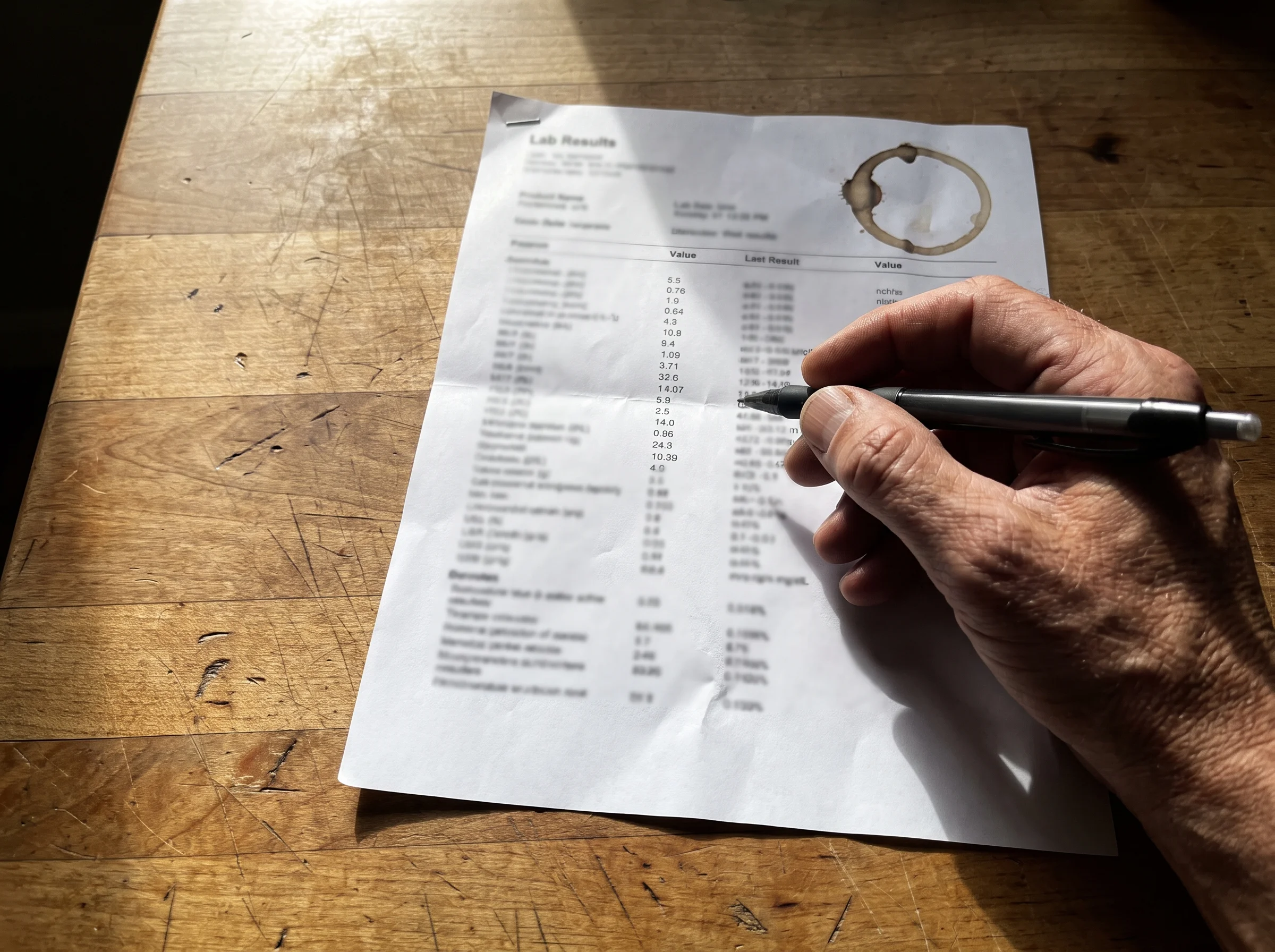 A close-up of a printed lab results sheet on a wooden kitchen table, coffee ring stain on one corner. A man's hand holding a 
