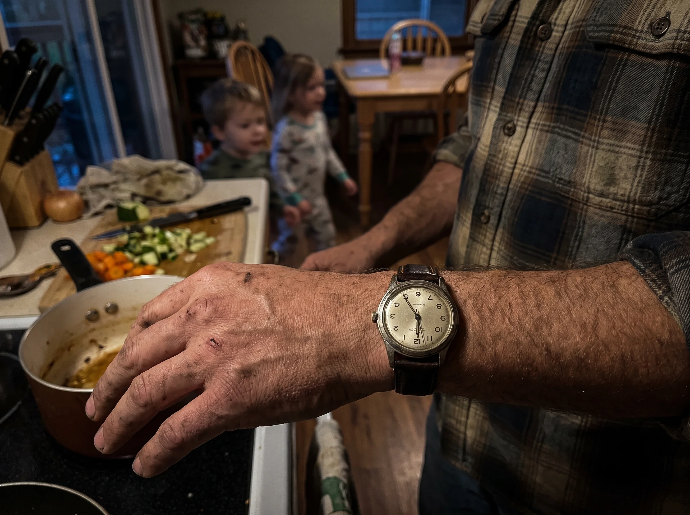 A close-up of a wristwatch reading 5:47pm on a tired man's arm as he stands in a suburban kitchen with dinner half-made, kids