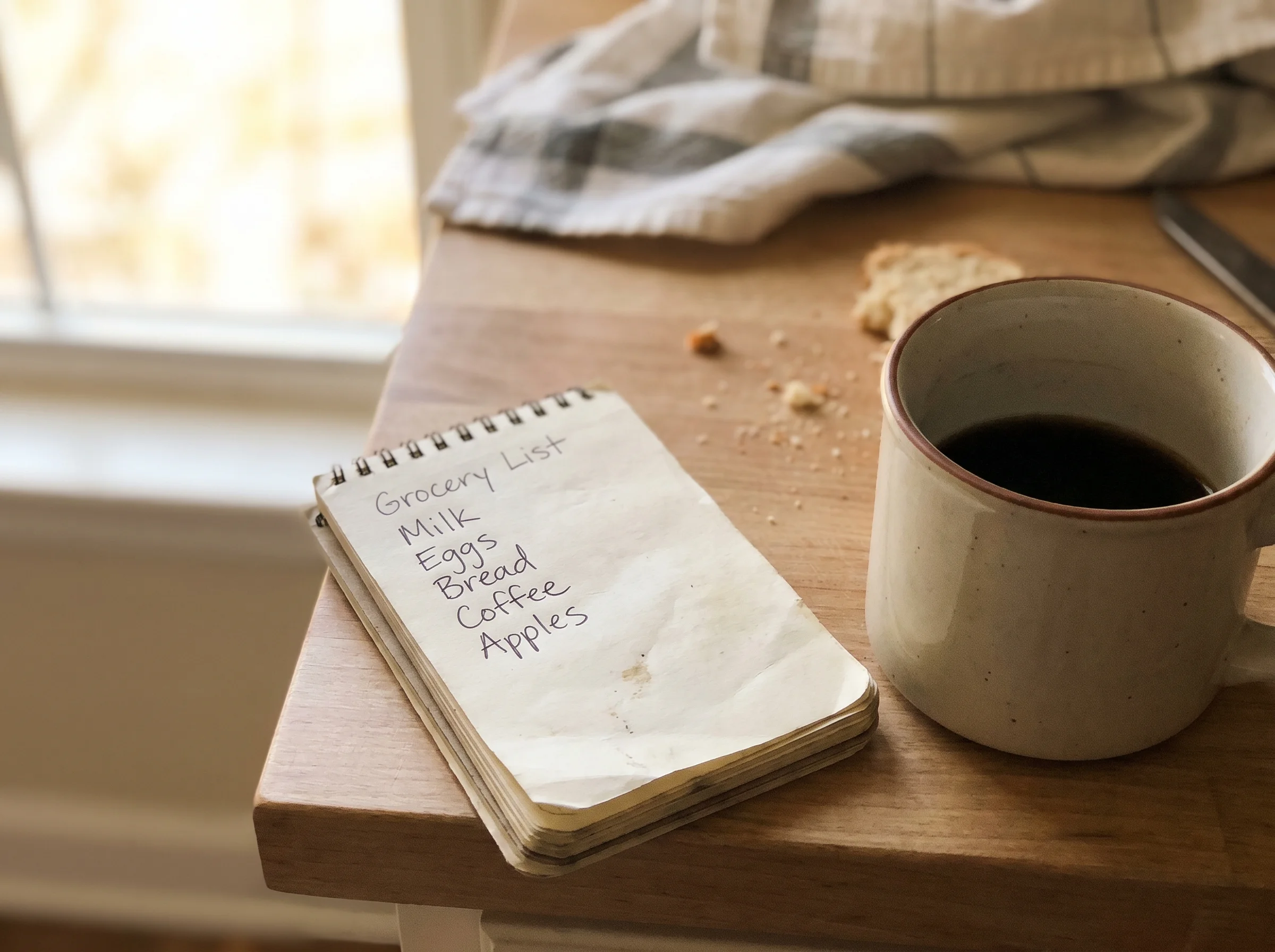 A handwritten list on a kitchen notepad next to a coffee mug, morning light, casual and slightly out of focus, shot on iPhone