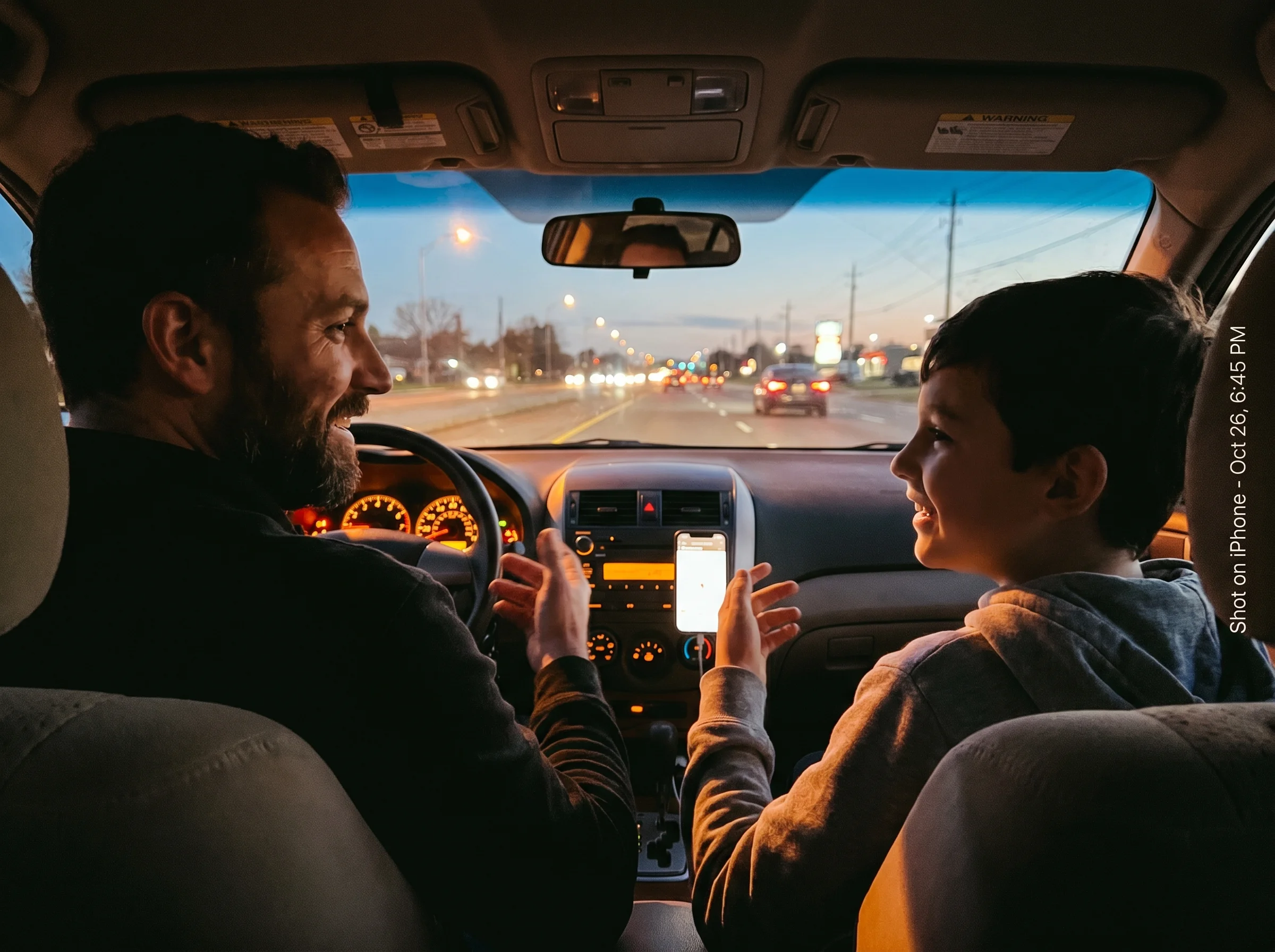 A dad and son talking in the front seats of a car at dusk, kid in the passenger seat, both relaxed and mid-conversation, warm