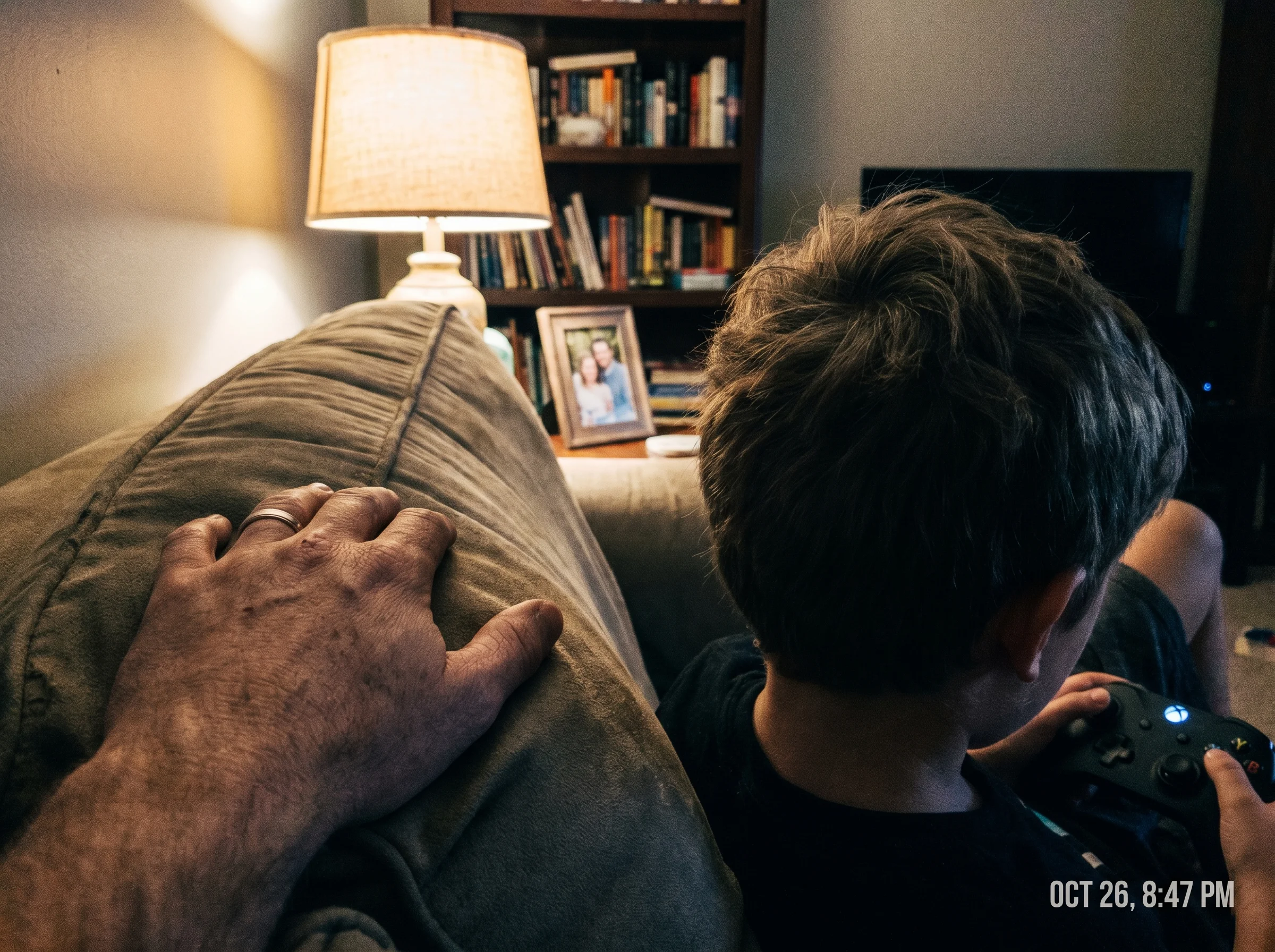 A close-up of a parent's hand resting casually on the back of the couch next to a kid focused on a game controller, soft lamp