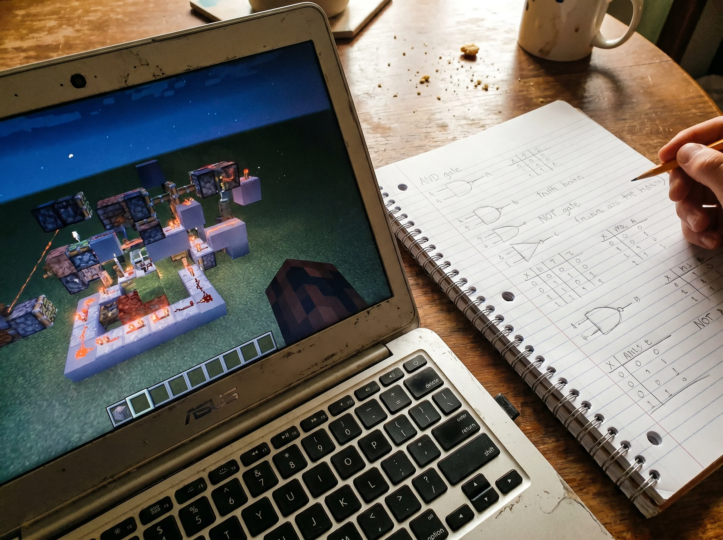 Close-up of a laptop screen showing a Minecraft redstone circuit, a notebook with hand-drawn binary logic sketches beside it