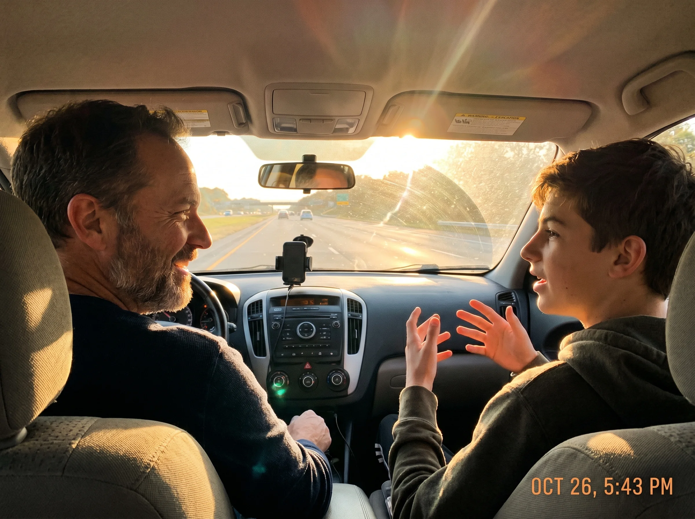 A dad and teen son in a car, son in passenger seat mid-conversation with hands gesturing, dad smiling at the road, late after