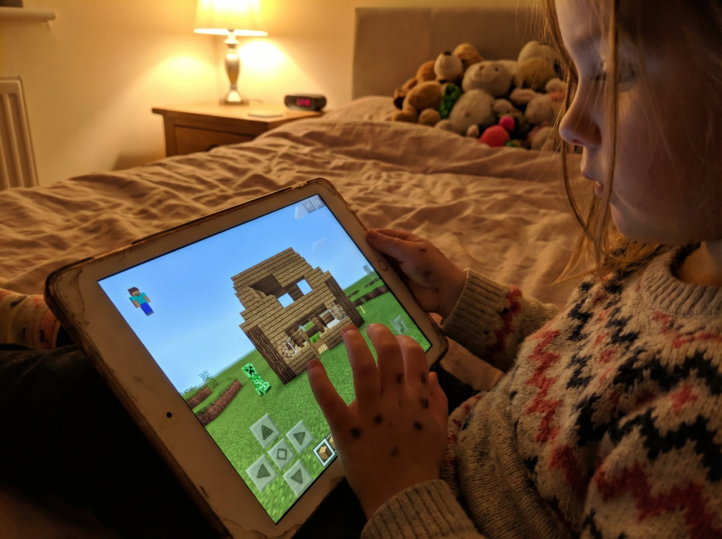 A young girl's hands gripping a tablet tightly, her Minecraft world visible on screen showing a partially destroyed house, so