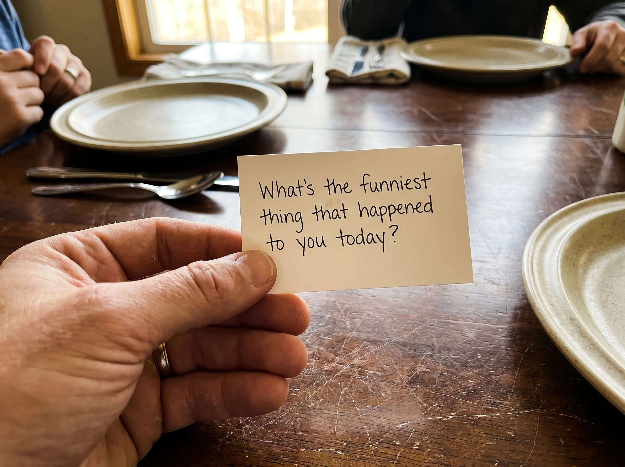Close-up of a hand holding a single conversation card above a worn wooden dinner table, dinner plates slightly out of focus i