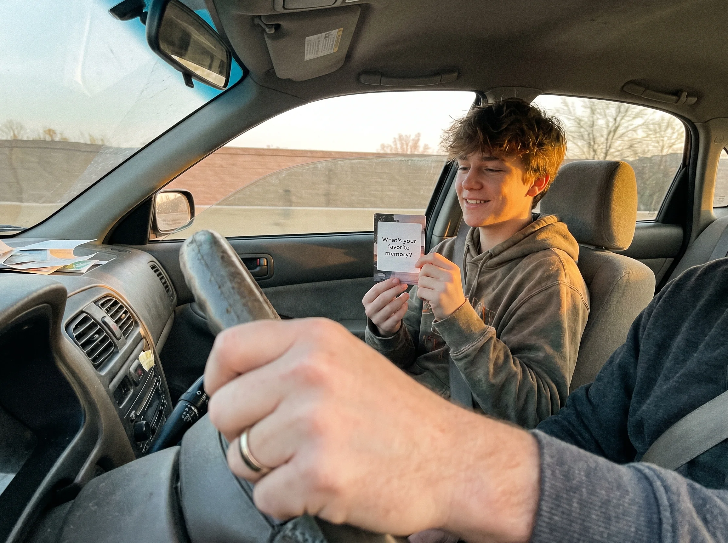 A teenage boy in the passenger seat of a car holding a conversation card, sunlight catching the side of his face, dad's hand