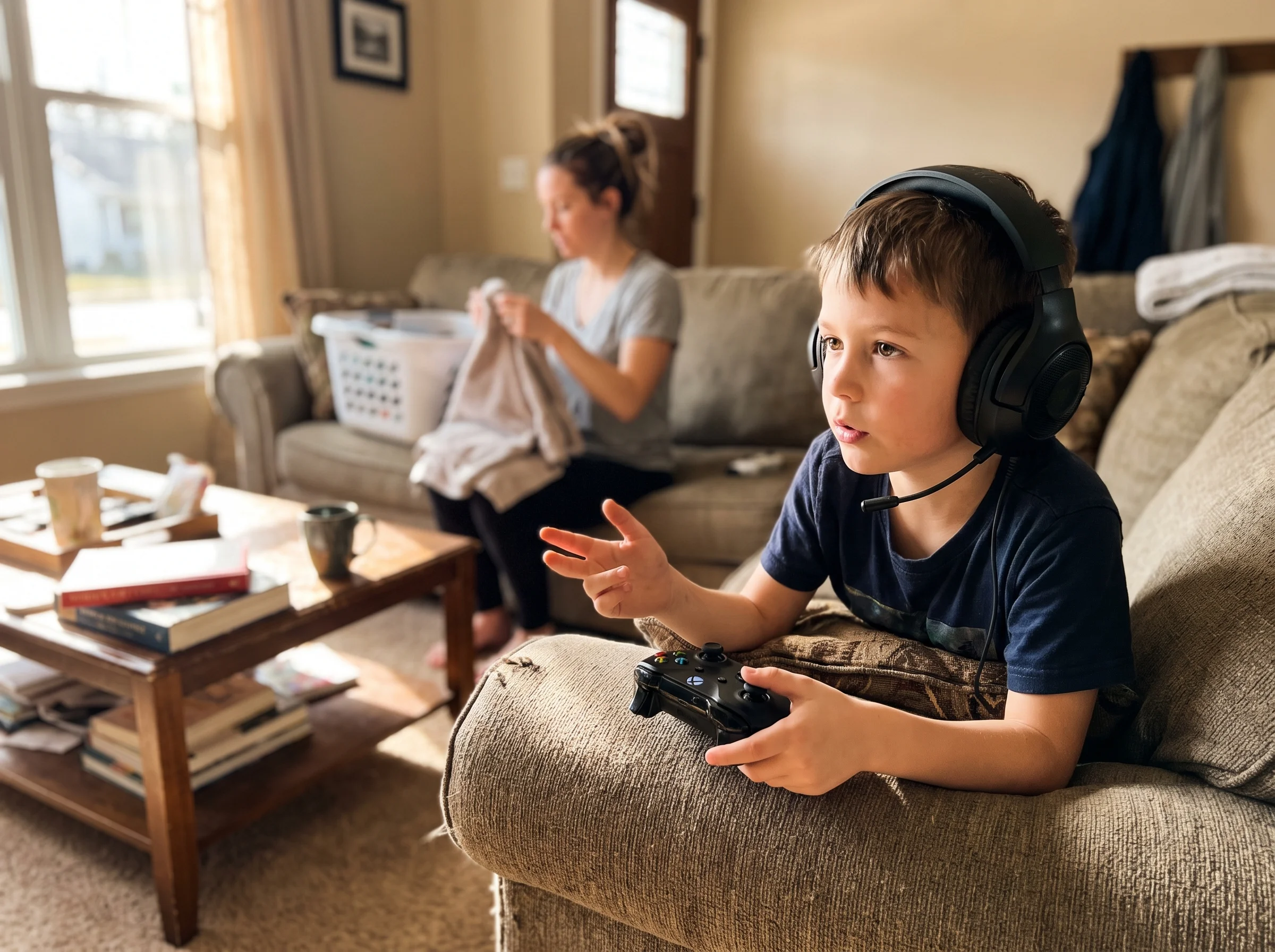 A kid wearing a gaming headset, mid-conversation with online squadmates, focused expression, controller in hand, mom in the b