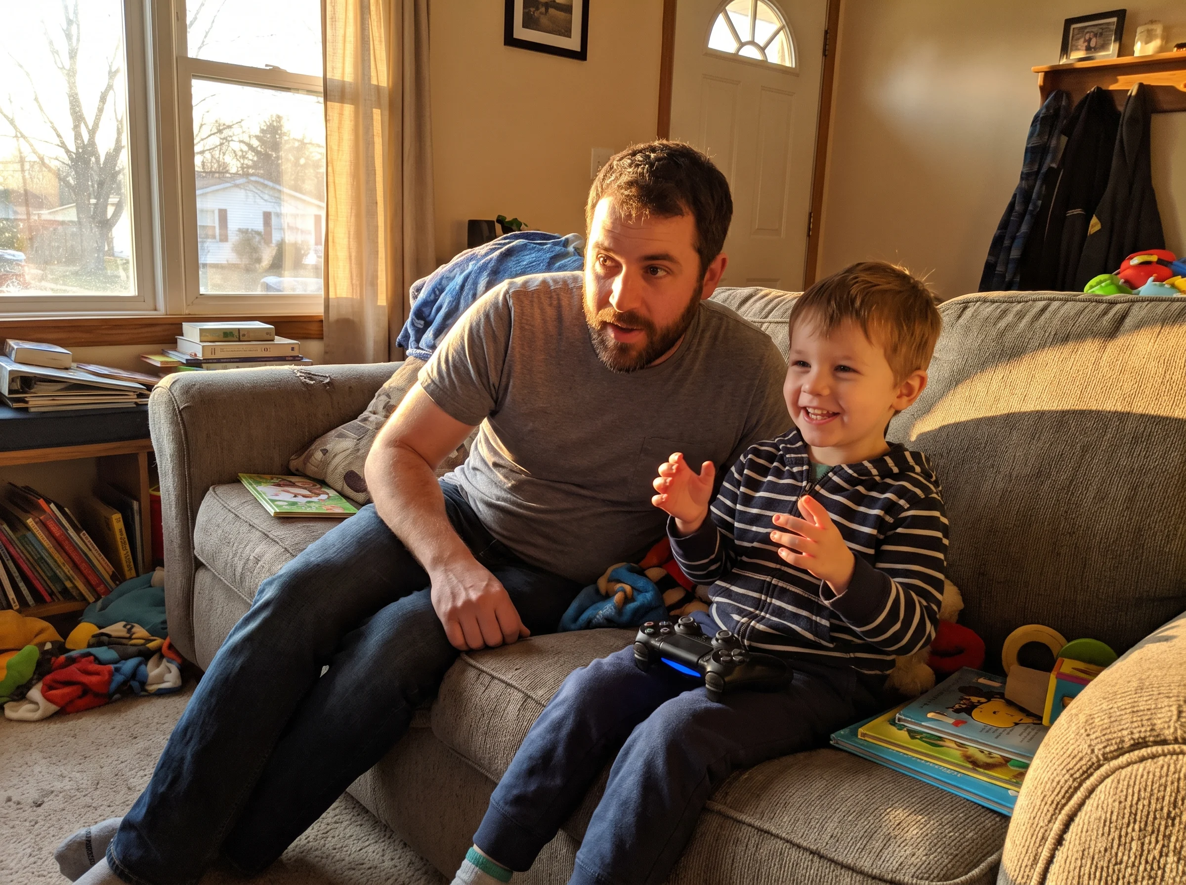 A father and son sitting on a couch, dad leaning in, son smiling and gesturing while telling a story, controller resting on h