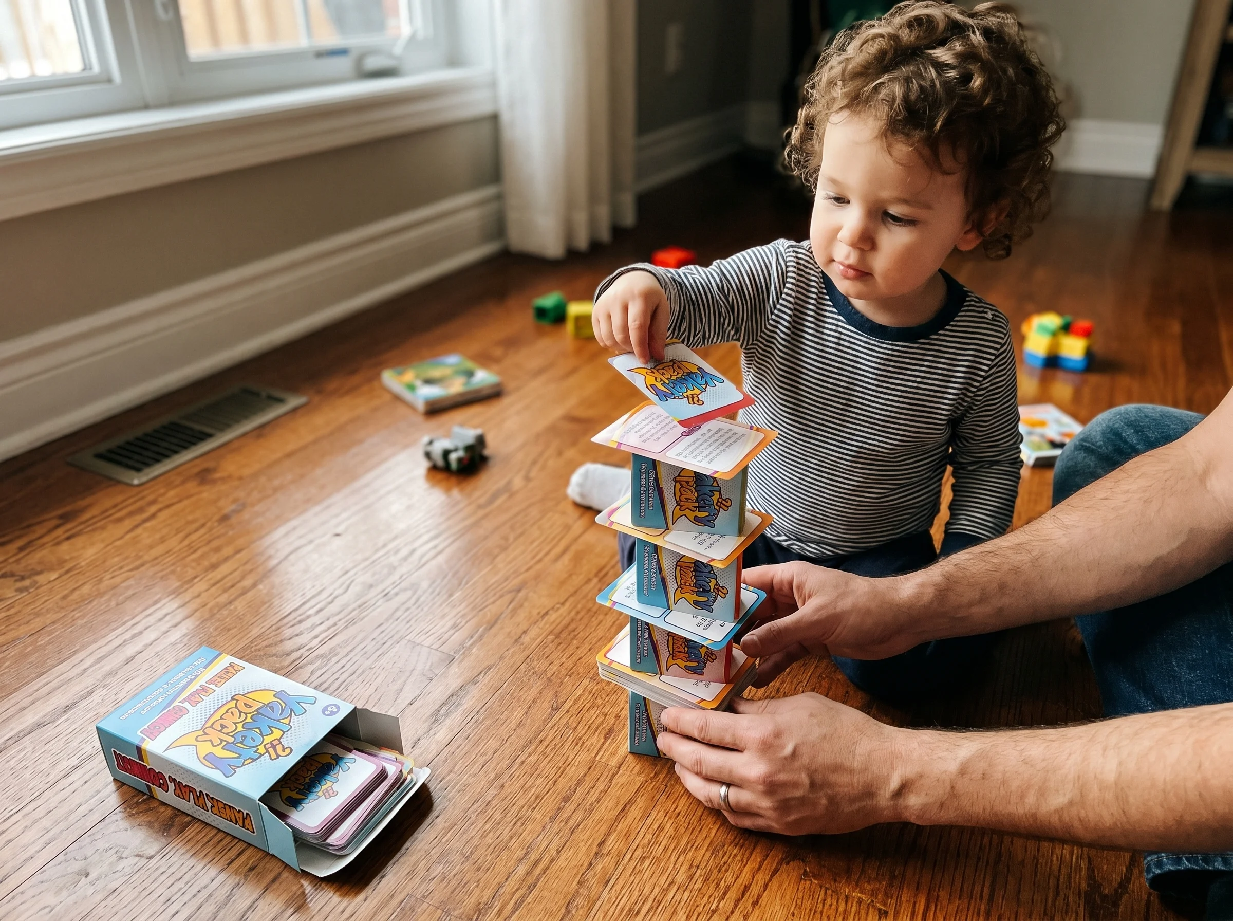 A toddler building a tall wobbly tower out of game cards on a hardwood floor, dad's hands visible steadying the base, natural