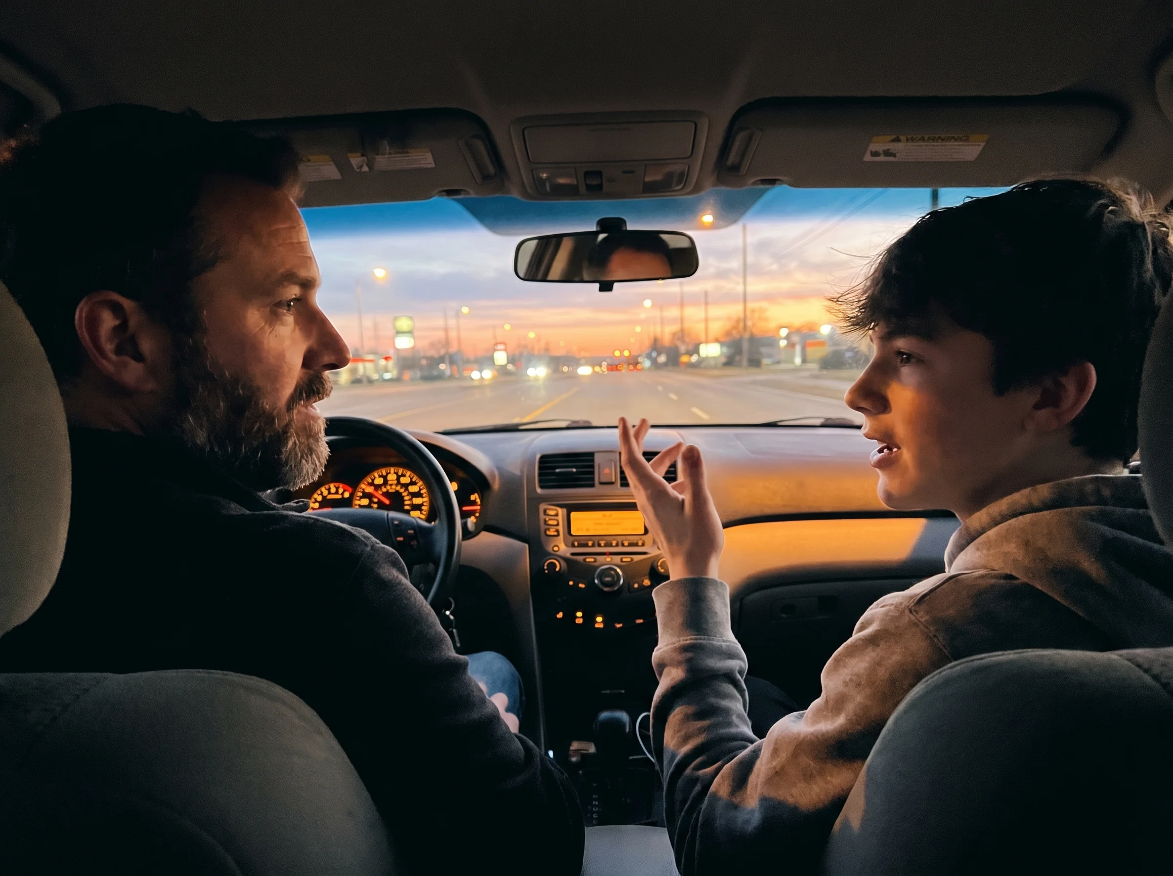 Dad and teenager in a car at dusk, dad driving, teen in passenger seat talking with hands, warm dashboard glow, casual conver