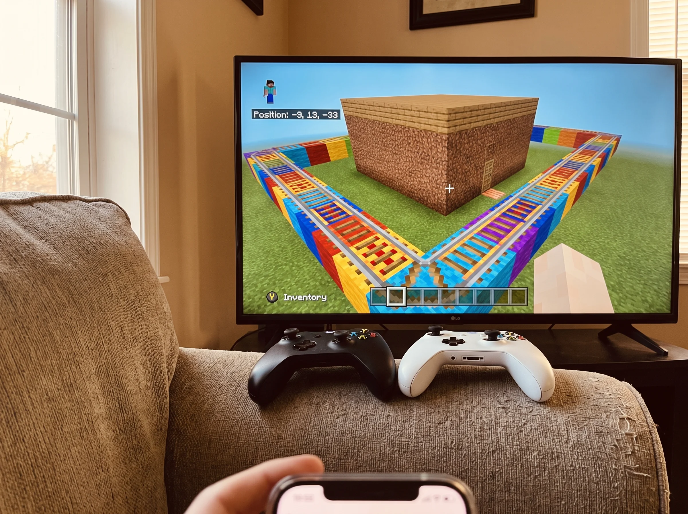 Close-up of a Minecraft build on a TV screen showing a colorful roller coaster next to a plain block house, with two controll