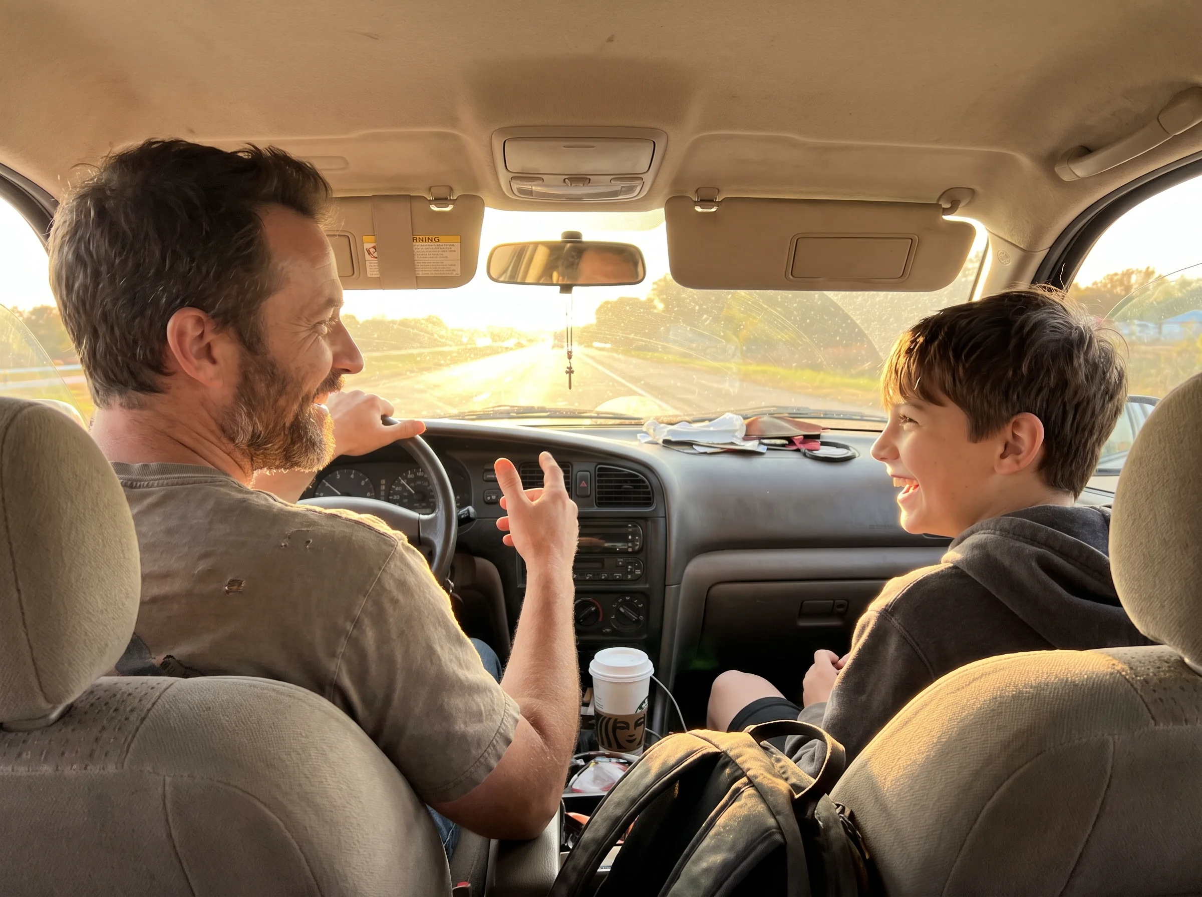 A dad driving with his son in the passenger seat, both mid-conversation, late afternoon light coming through the windshield