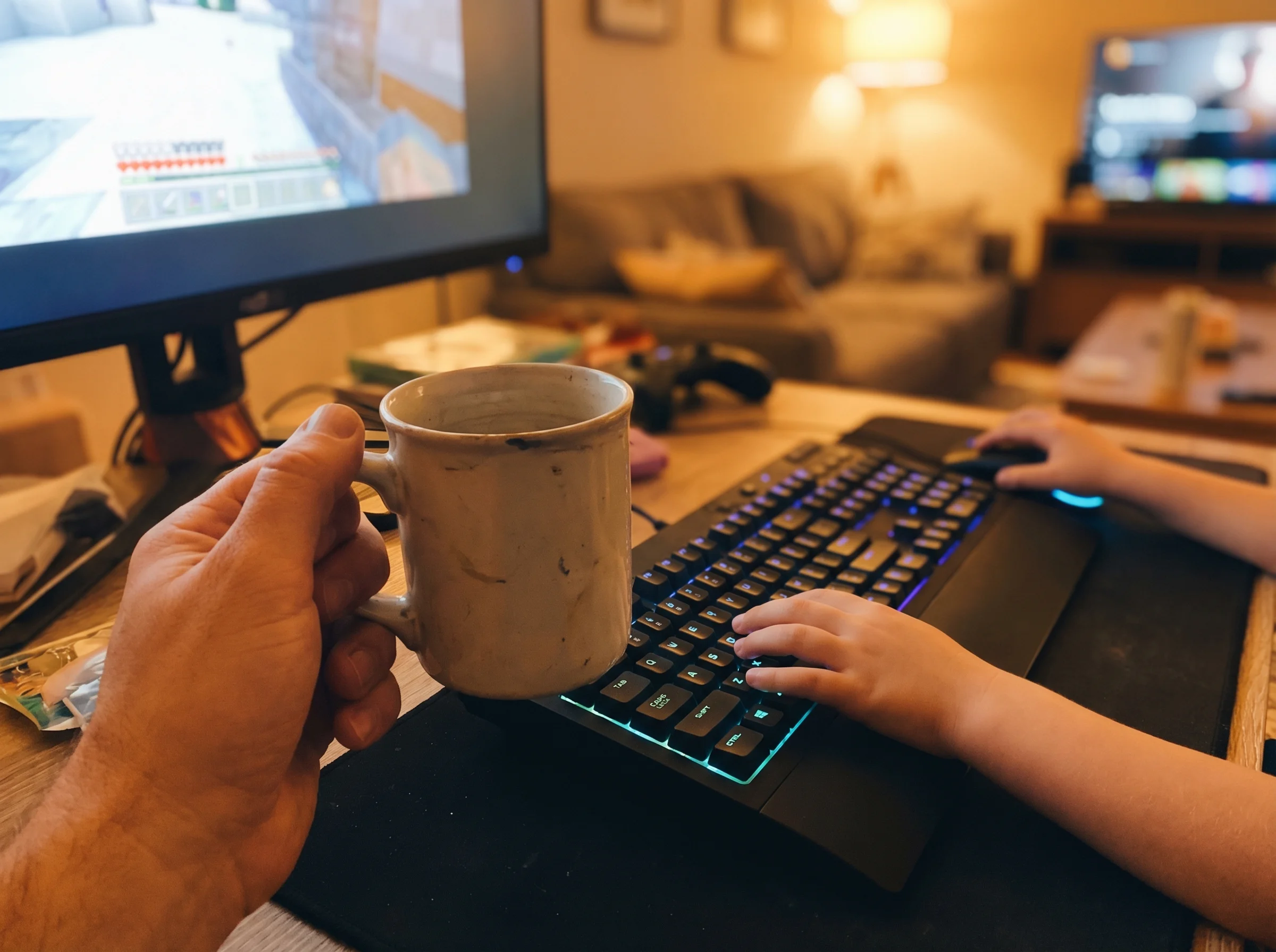 Close-up of dad's hand holding a mug while sitting next to gaming setup, kid's hands visible on keyboard and mouse, warm even