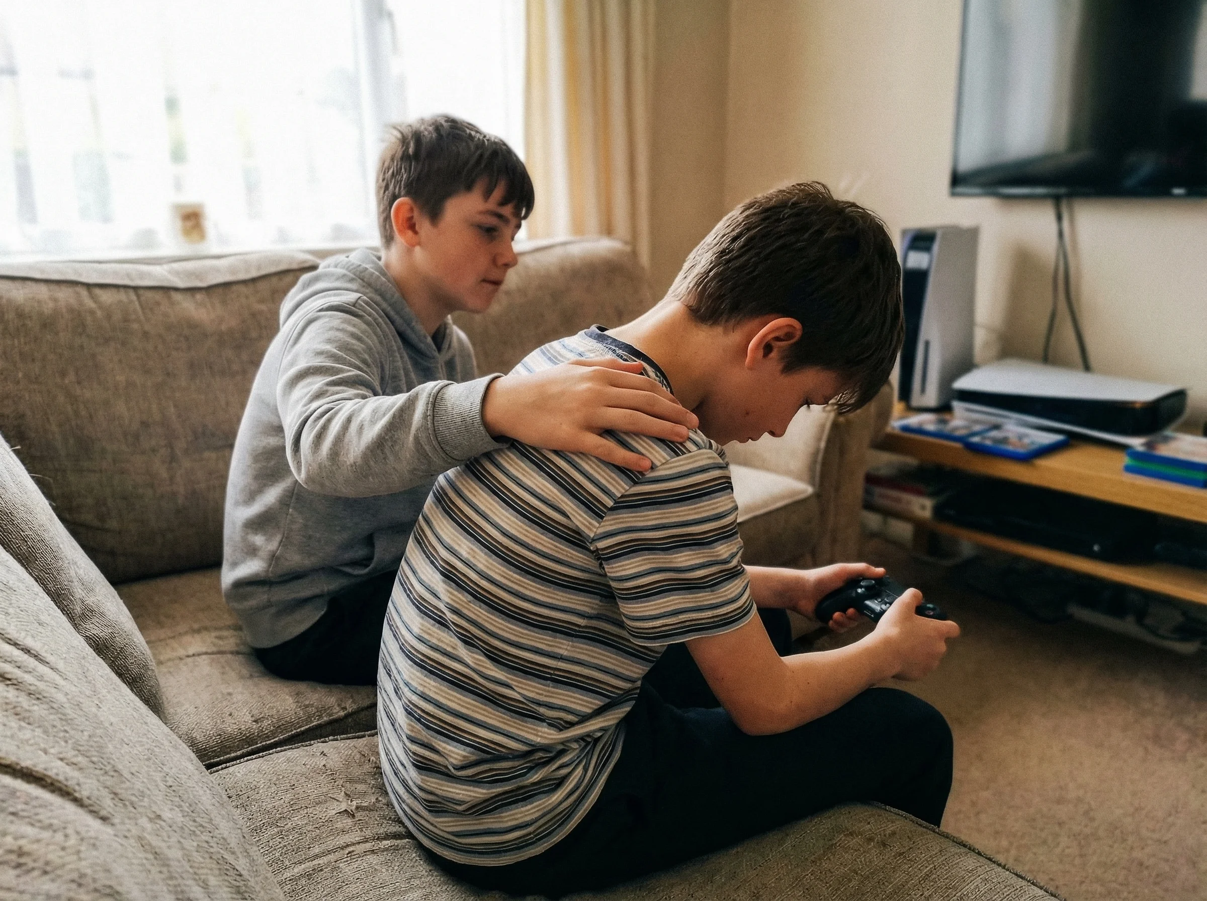Two boys around 13 years old sitting on a couch, one consoling the other who looks upset while holding a gaming controller, P