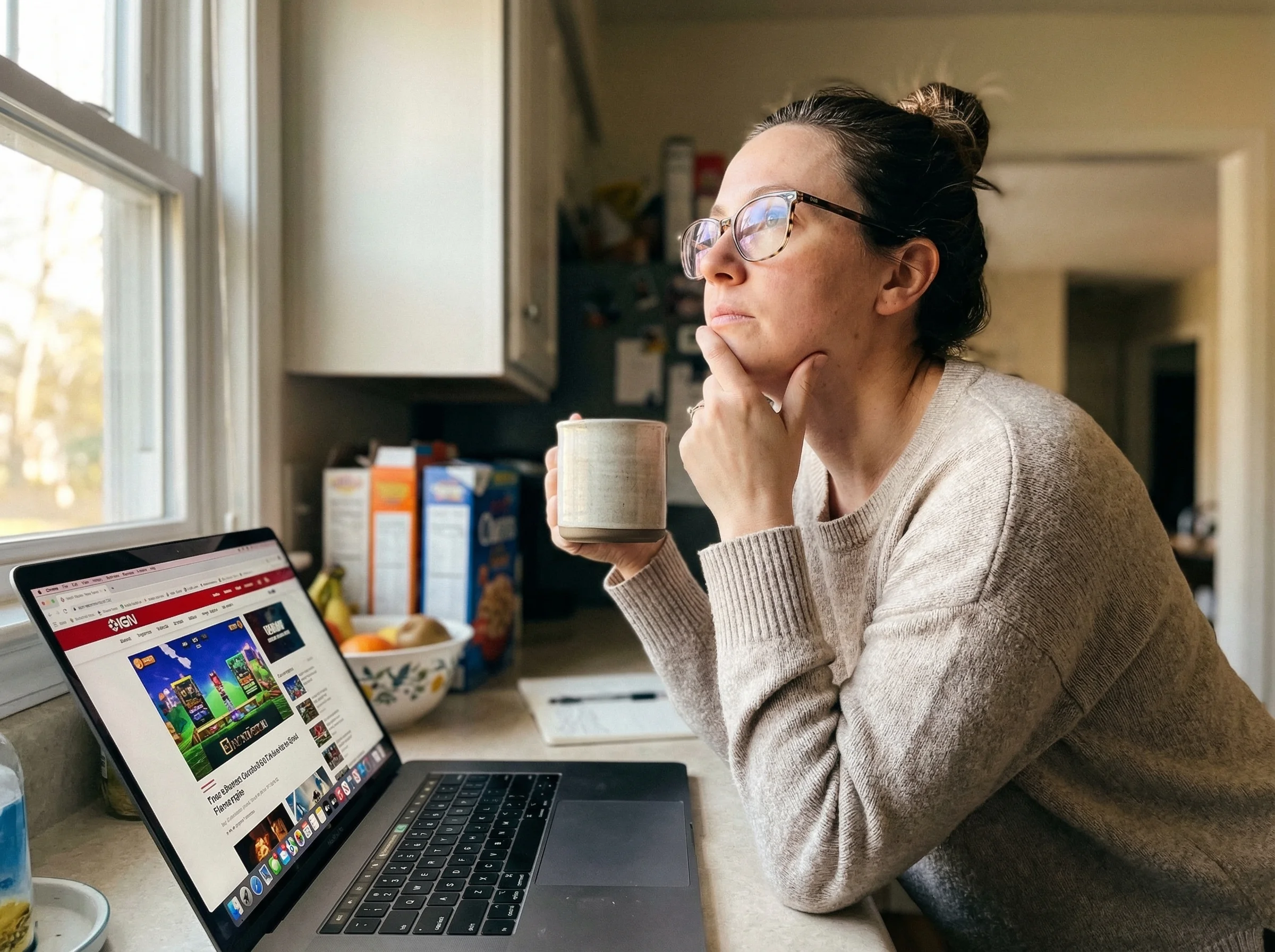 Mom sitting at kitchen counter with laptop open to a gaming website, looking thoughtful while researching, cup of coffee besi