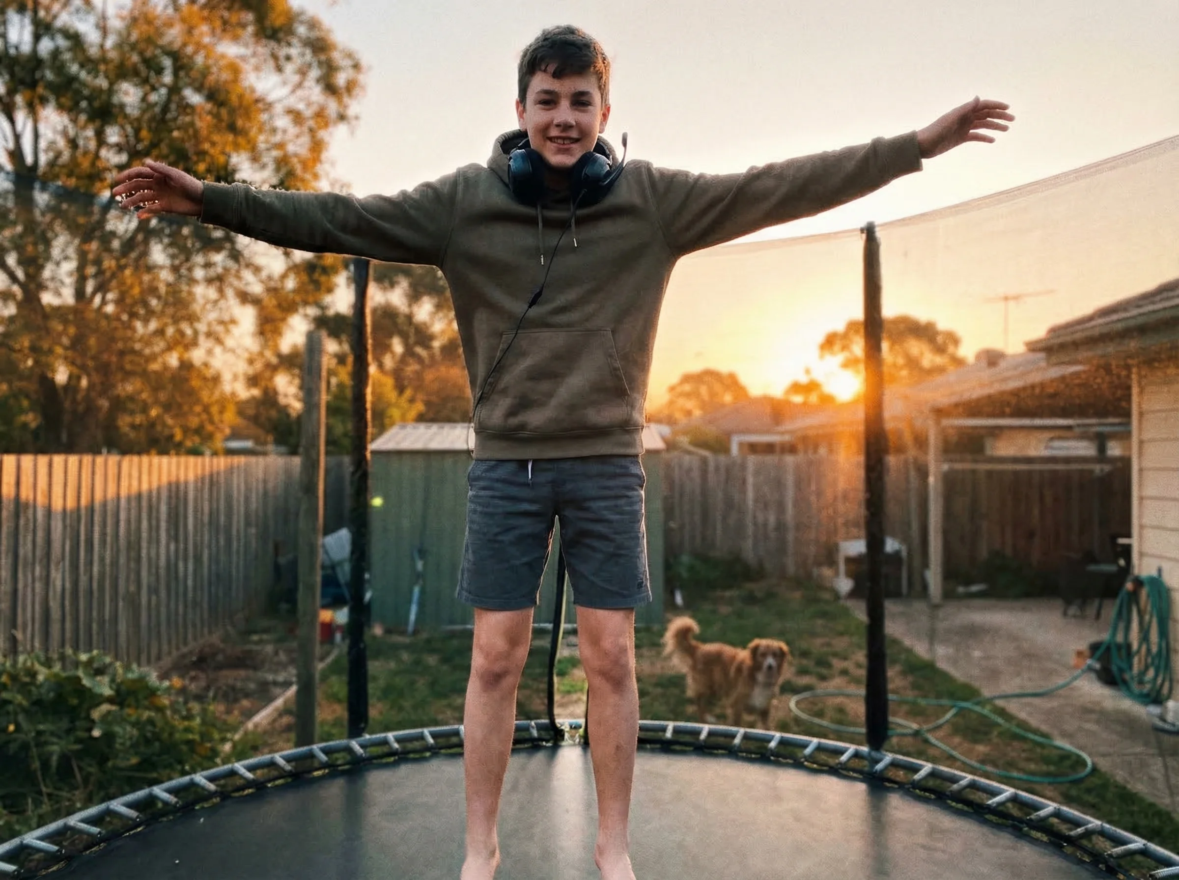 Teen boy on a trampoline in the backyard, mid-jump with arms spread wide, gaming headset still around his neck, golden hour l