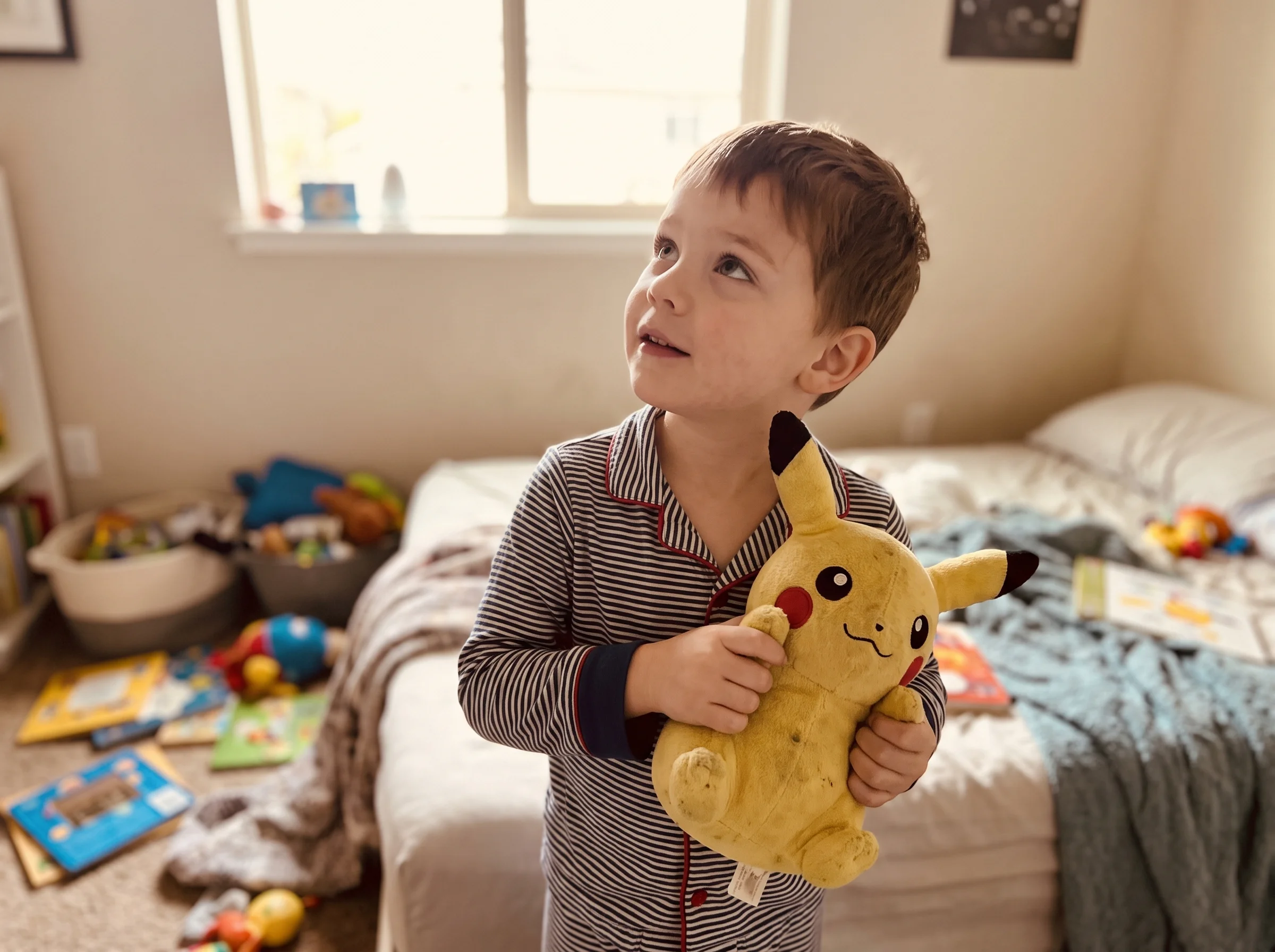 A young boy in pajamas holding a Pikachu plush, looking up curiously, casual bedroom setting, shot on iPhone with natural war