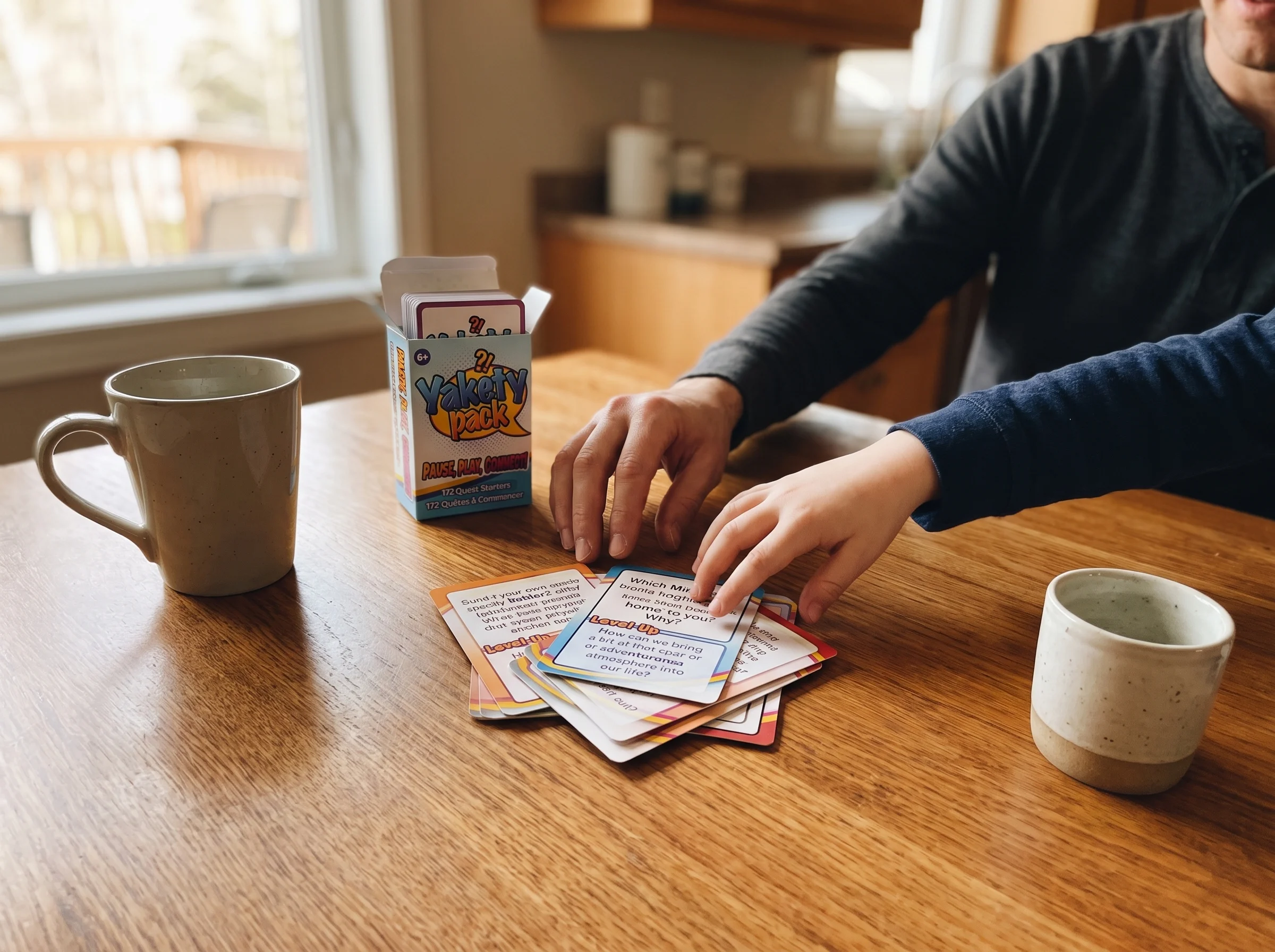 A small pile of Pokémon trading cards spread on a kitchen table next to two mugs, one kid-sized hand and one adult hand mid-r