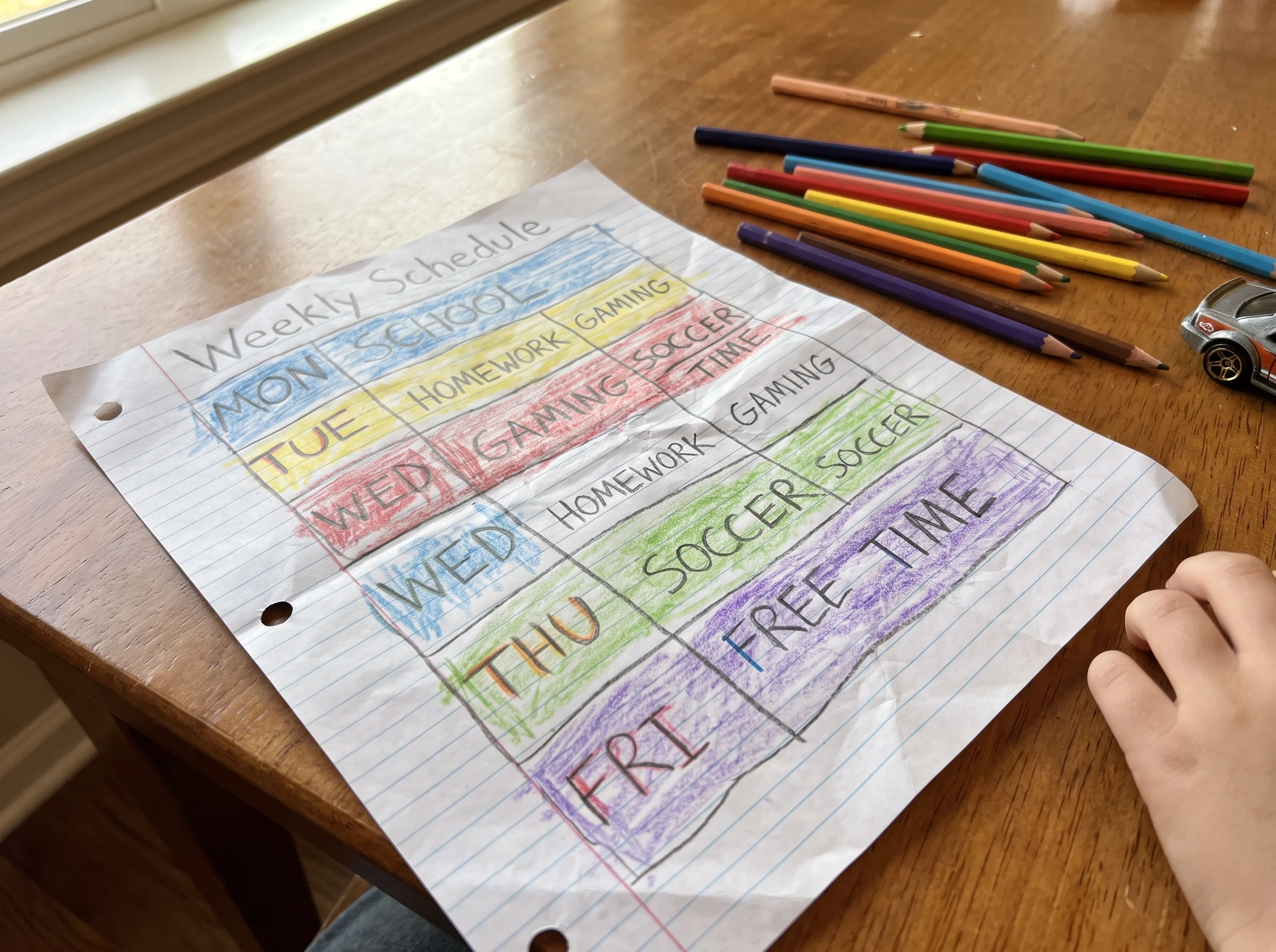 A handwritten weekly schedule on notebook paper with a kid's messy handwriting, colored pencils nearby, showing gaming and ho