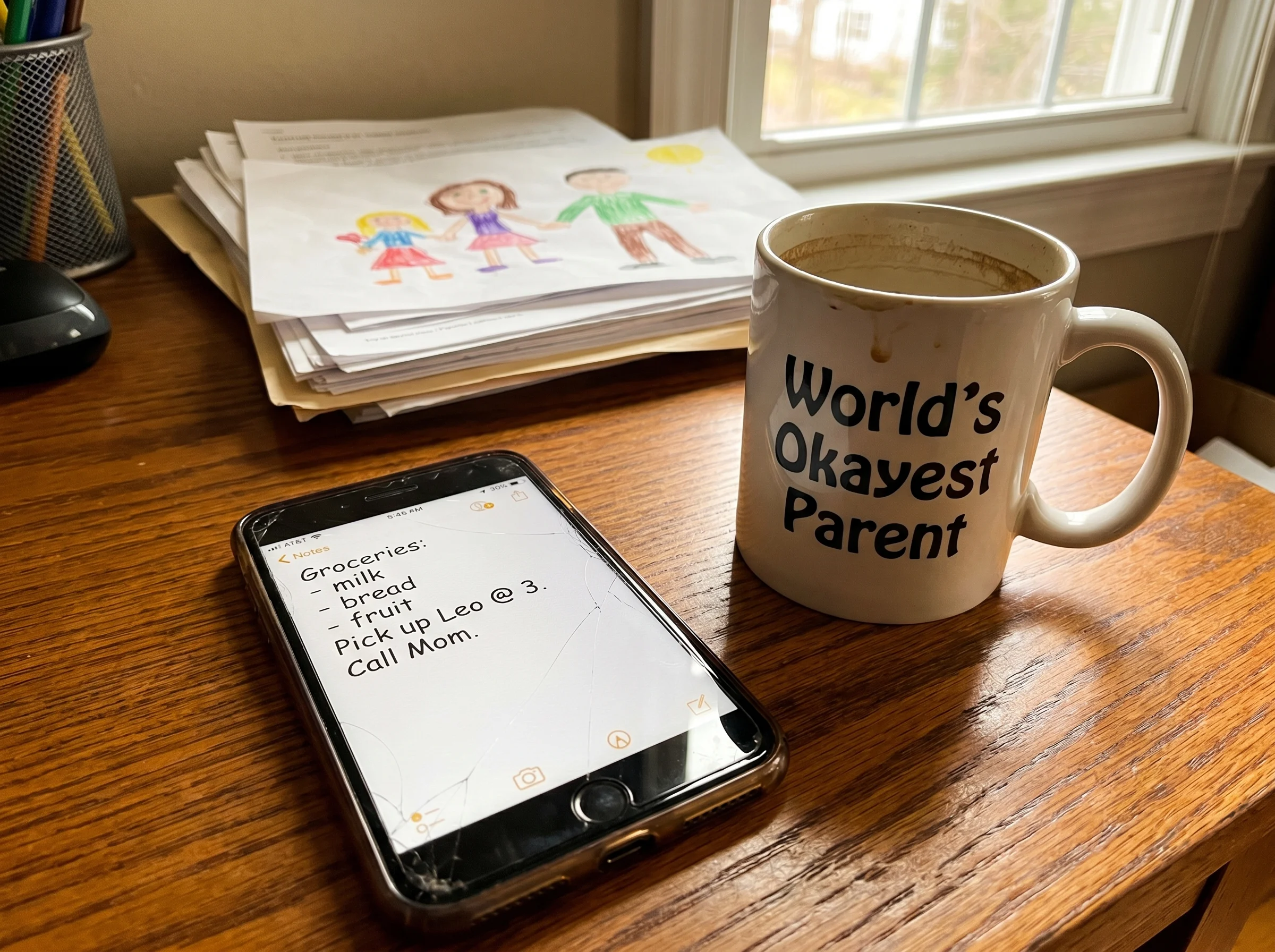 A parent's phone on a desk showing a simple notes app entry with a few short lines of text, coffee mug beside it, casual home