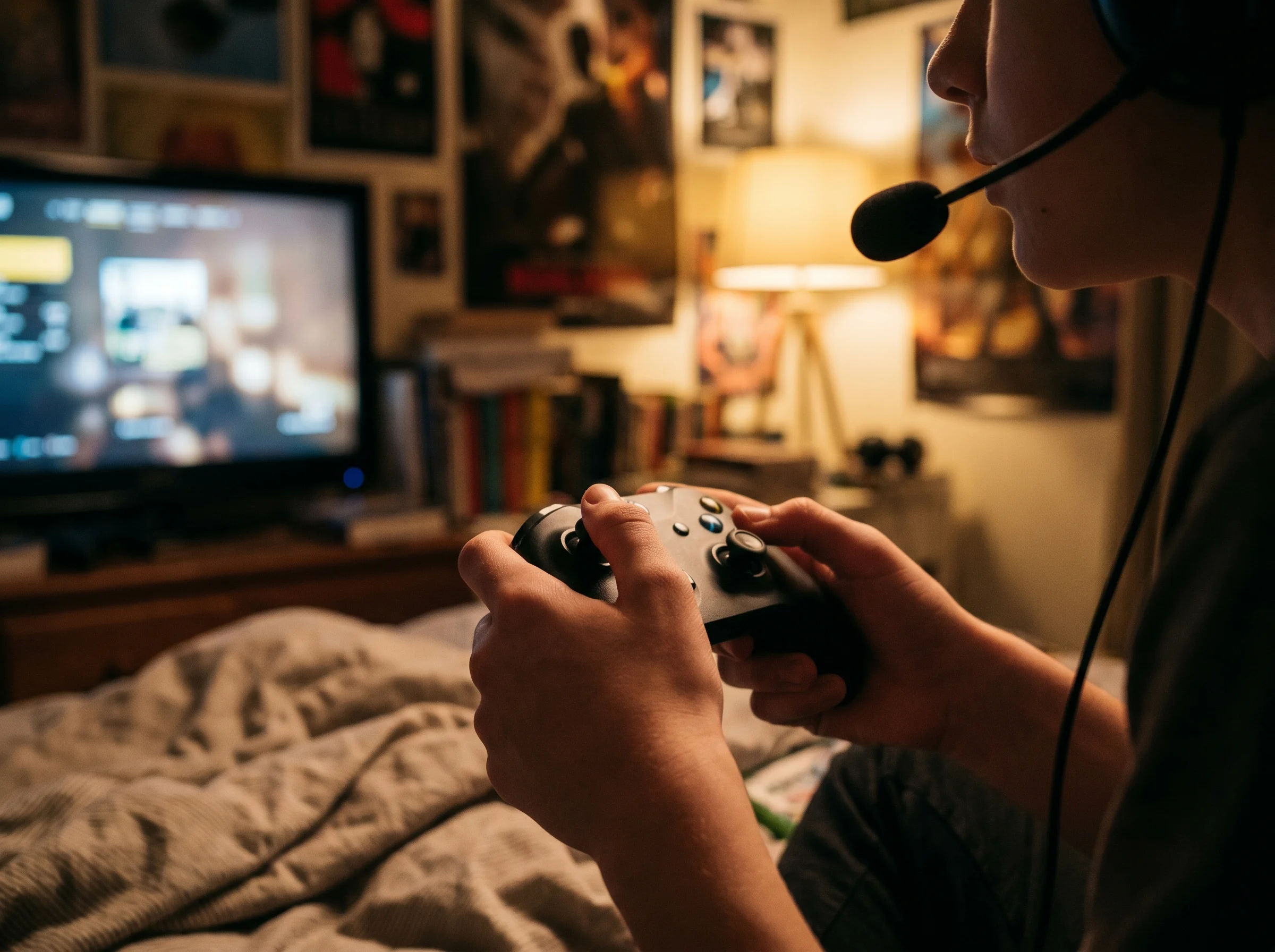 Close-up of a teen's hands on a game controller with a headset mic visible, screen glowing in the background, candid bedroom 