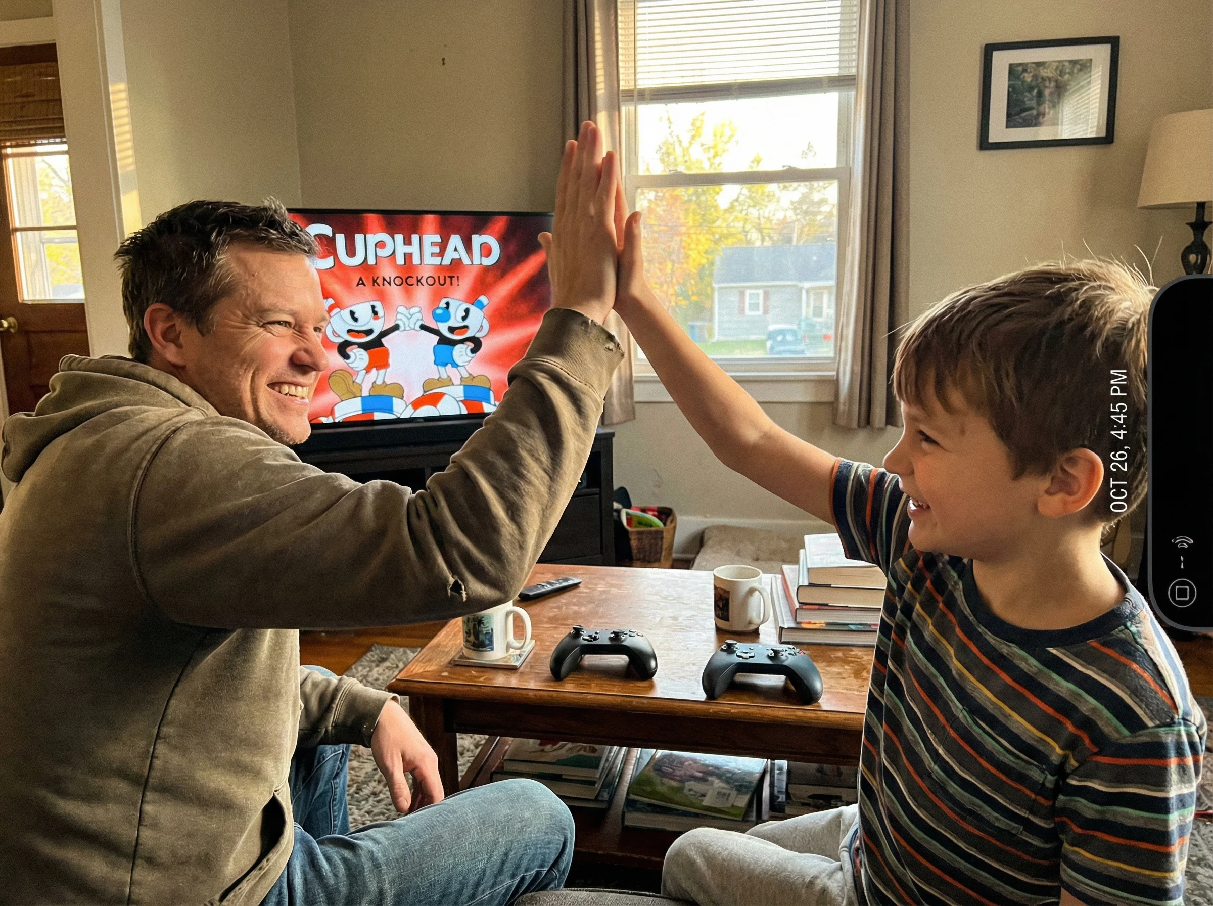 Parent and child high-fiving in front of TV showing Cuphead victory screen, game controllers on the coffee table, genuine smi