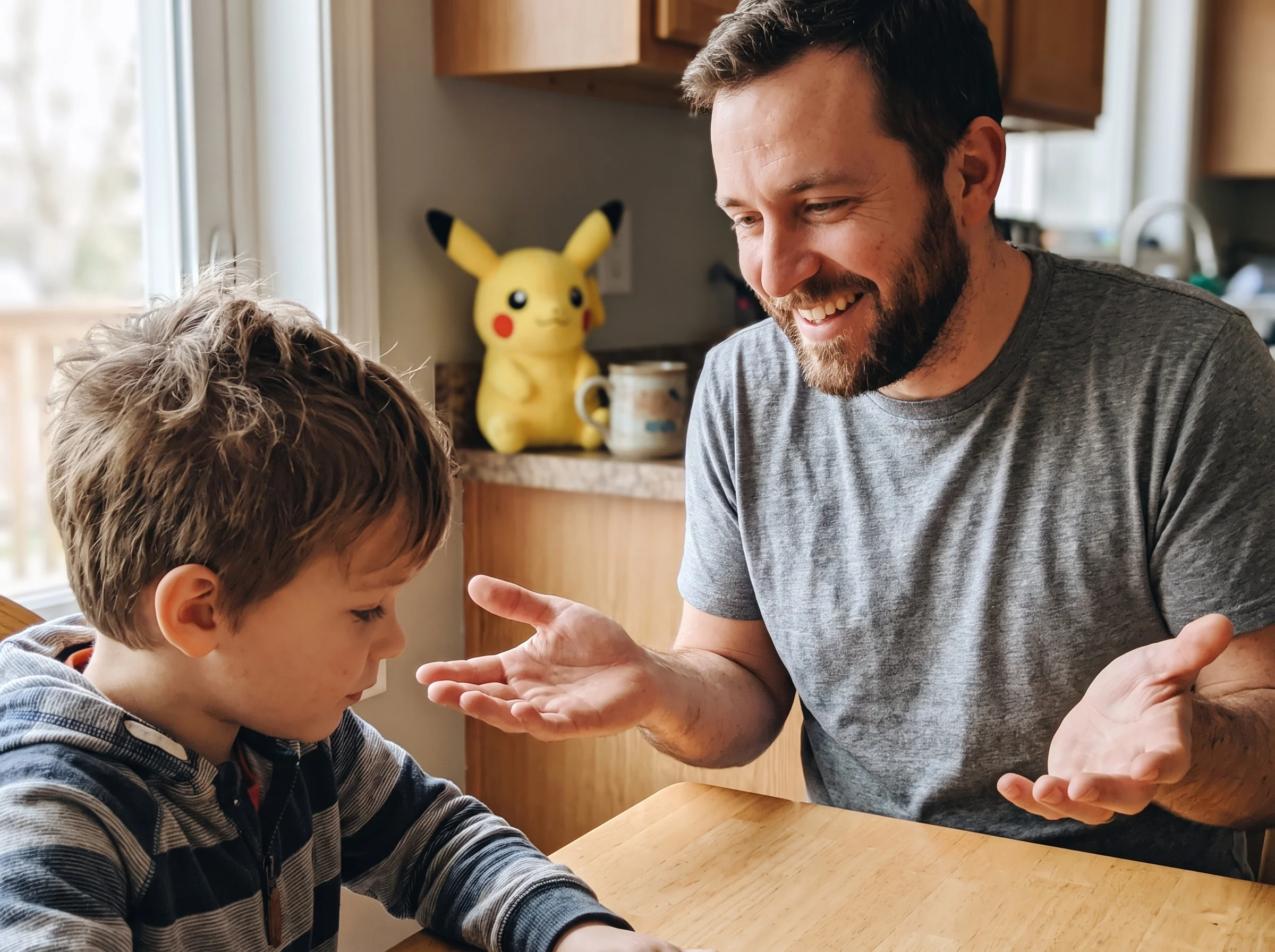 Close-up of a dad smiling and shrugging with hands open, sitting across from his son at a kitchen table, plush Pikachu in the