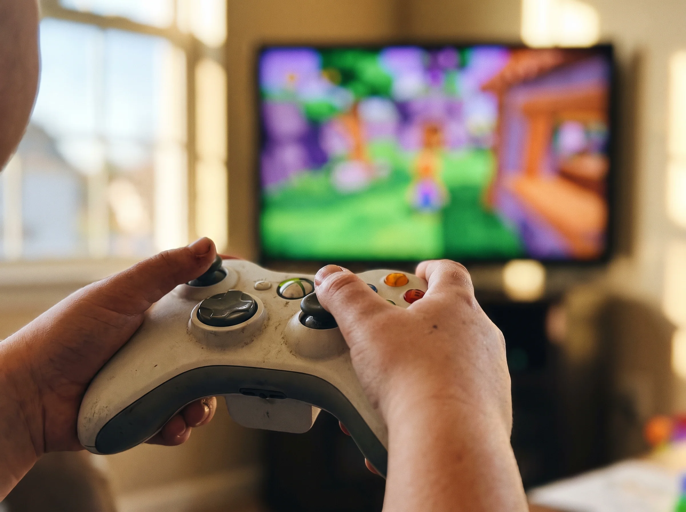 Close-up of a kid's hands holding a game controller, screen blurred in background showing colorful Fortnite-style graphics, n