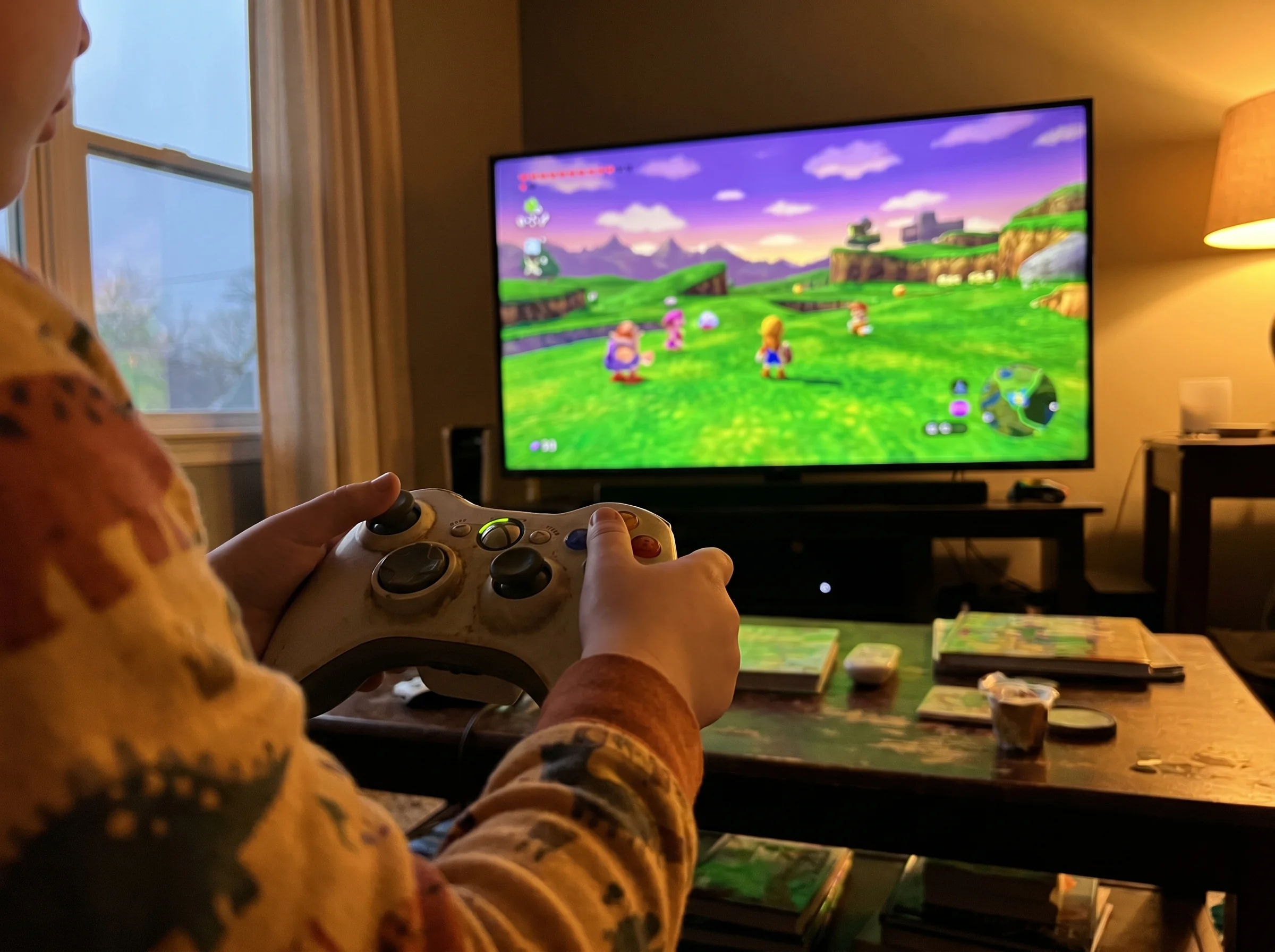 Close-up of a TV screen showing a colorful open-world game, with a kid's hands on a controller in the foreground, soft evenin