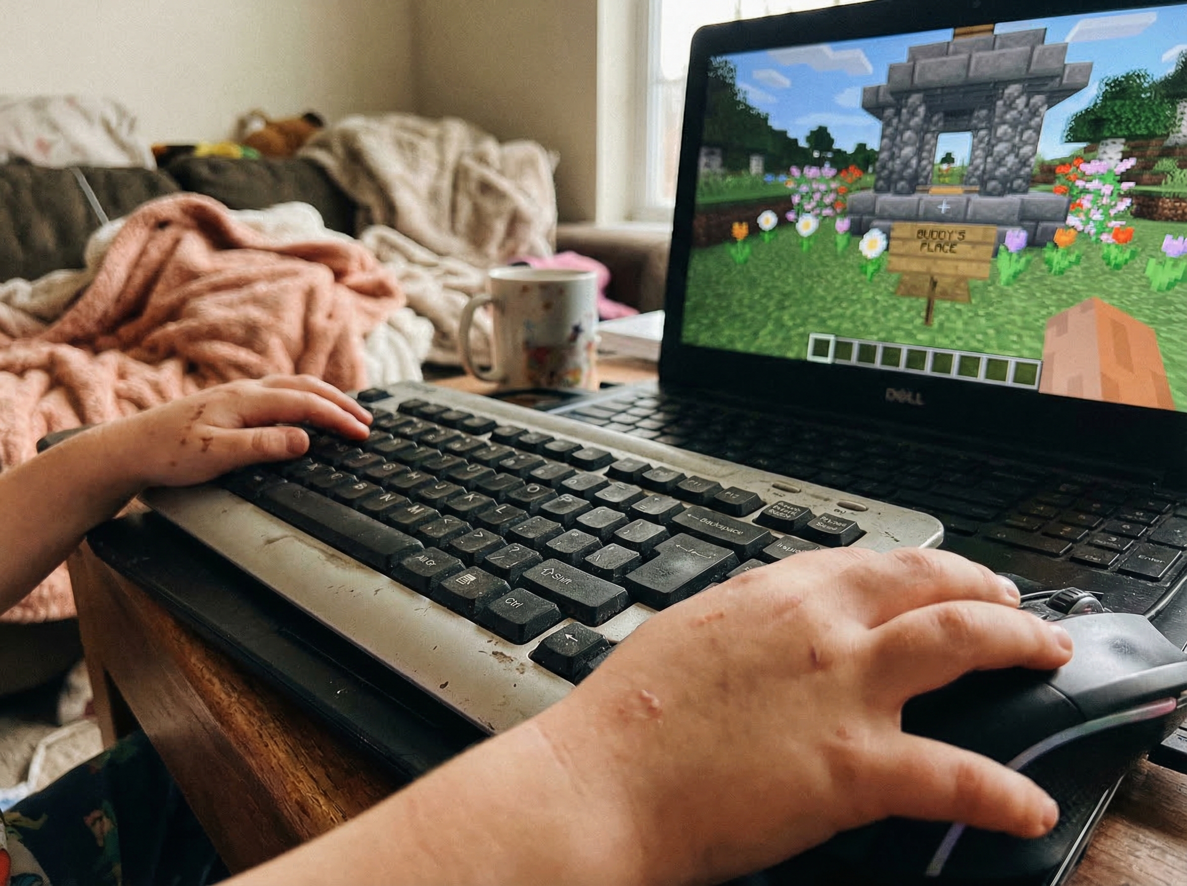 Close-up of a child's hands on keyboard and mouse, Minecraft memorial garden visible on screen with "Buddy's Place" sign clea