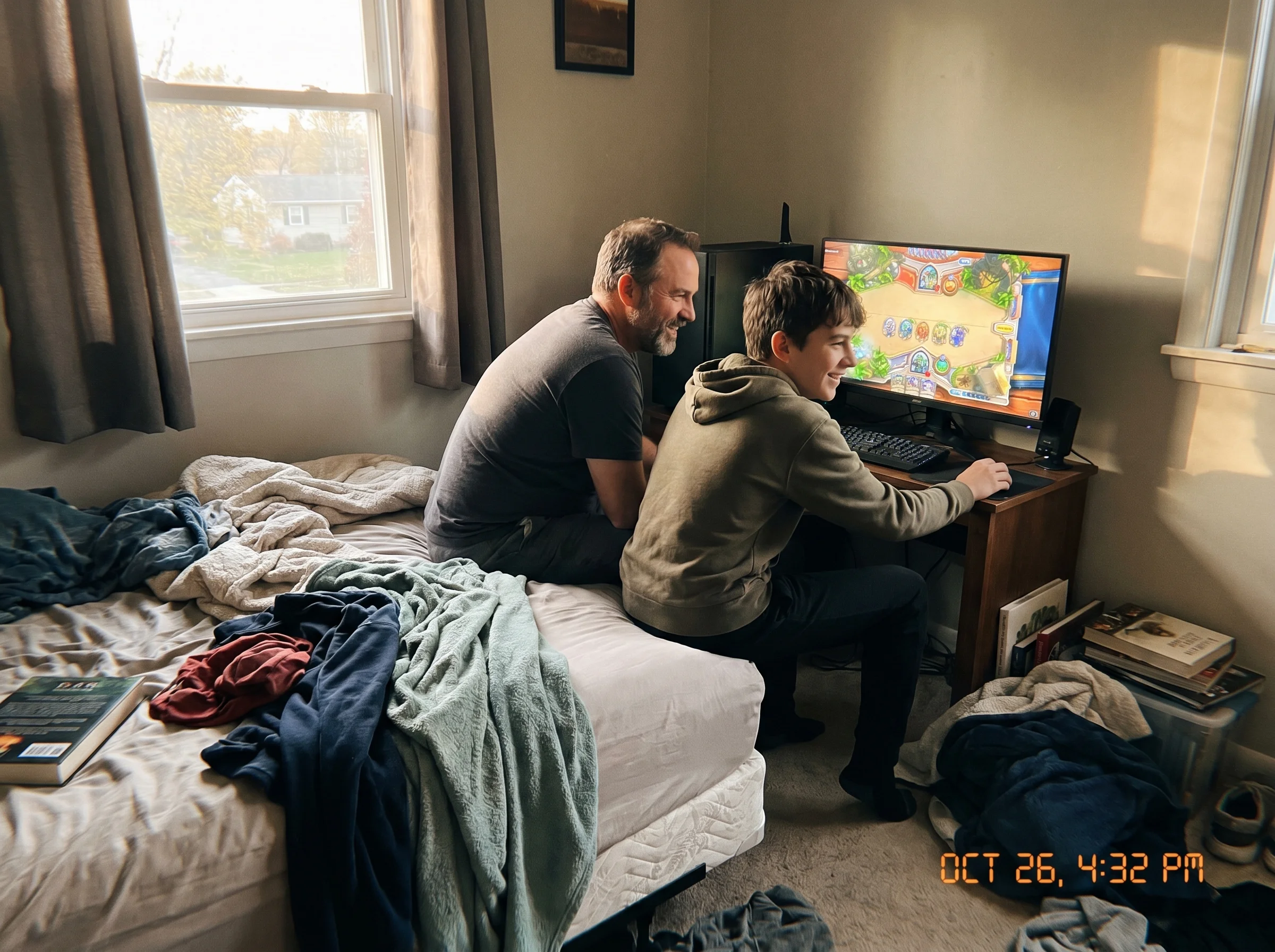 A dad and teen son sitting on the edge of a bed together, the teen showing dad something on a gaming monitor, both leaning in