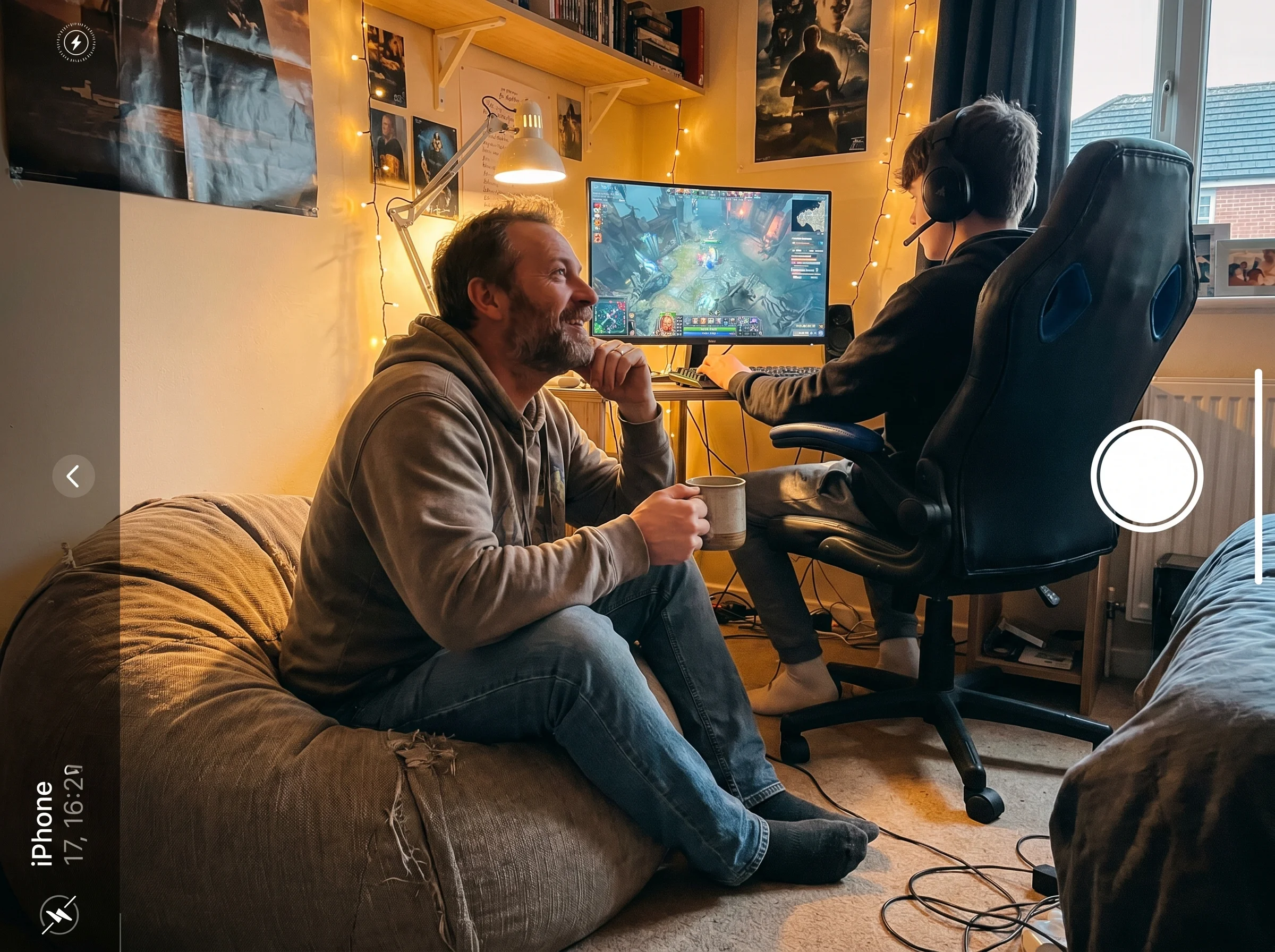 A dad sitting on a beanbag chair next to his teen's gaming setup, watching the screen with genuine curiosity, mug of coffee i