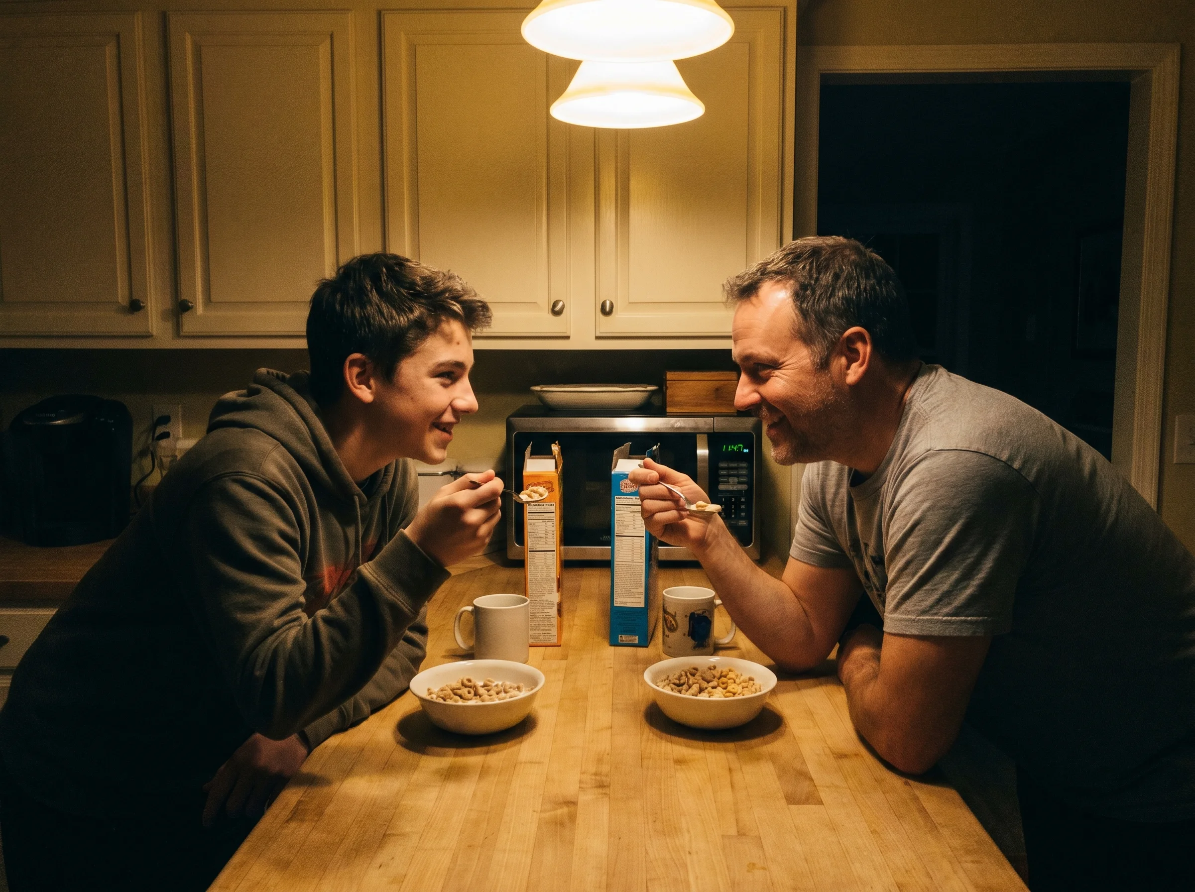 A teen boy and his dad at the kitchen counter eating cereal late at night, both relaxed, mid-conversation, shot on iPhone wit