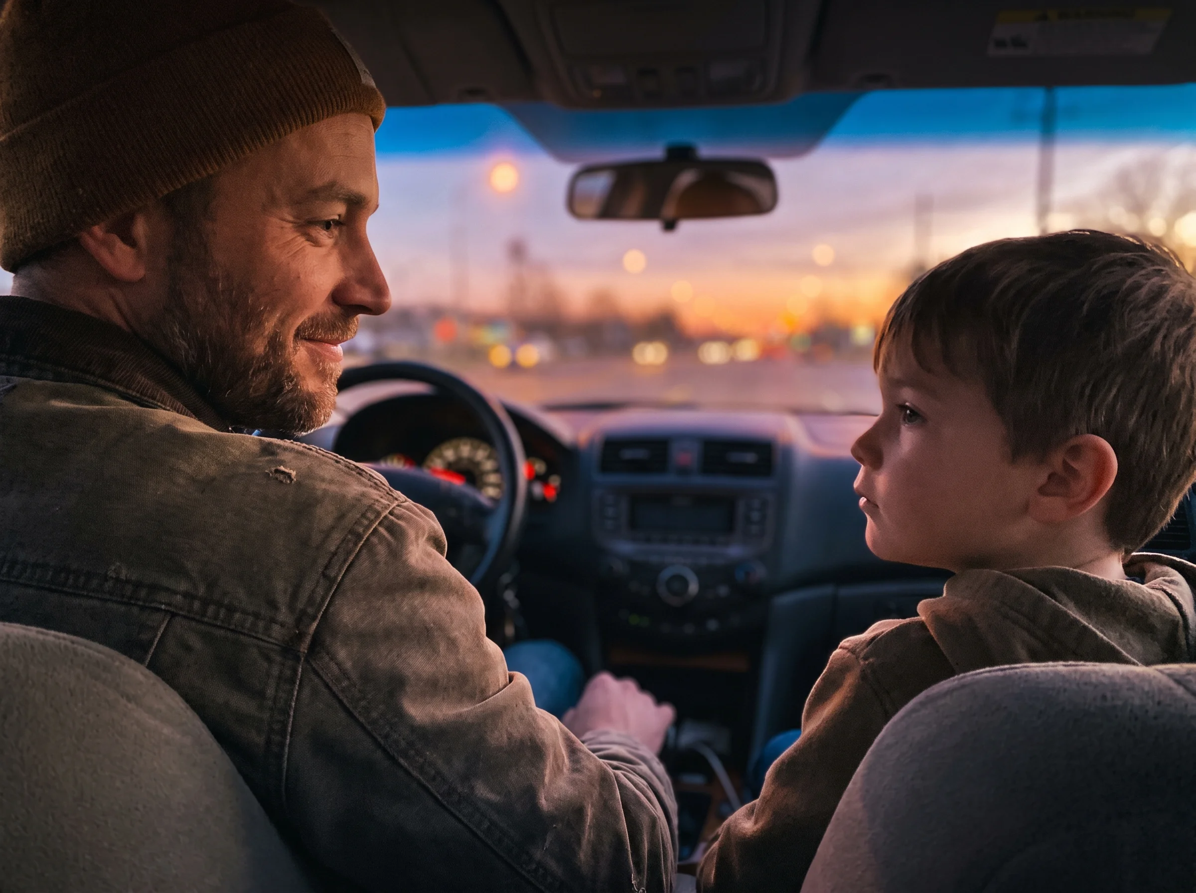 Father and son in the front seats of a car at dusk, son looking out the window thinking, dad glancing over with a small smile