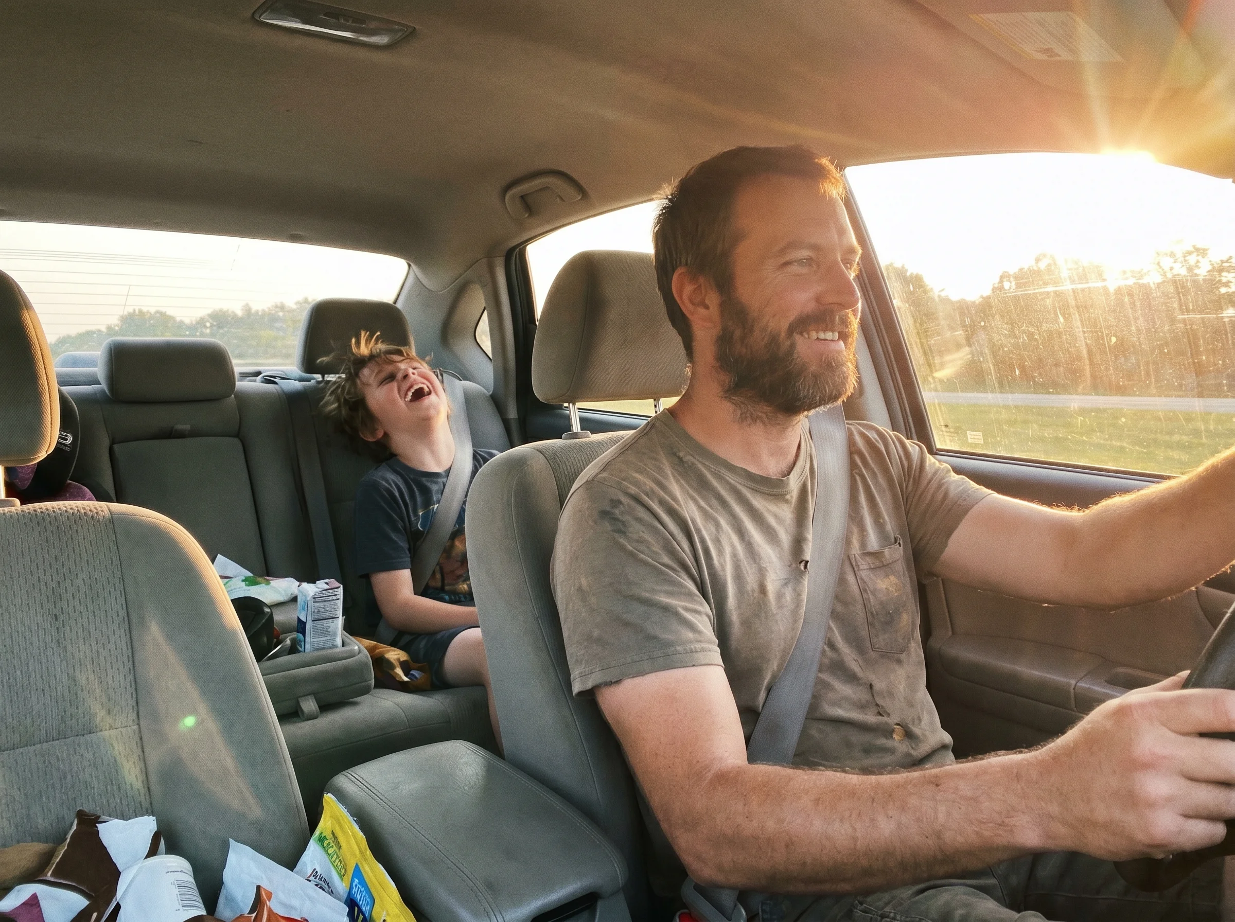 A view from the passenger seat of a car showing a dad driving and a kid in the back seat laughing, late afternoon sunlight th