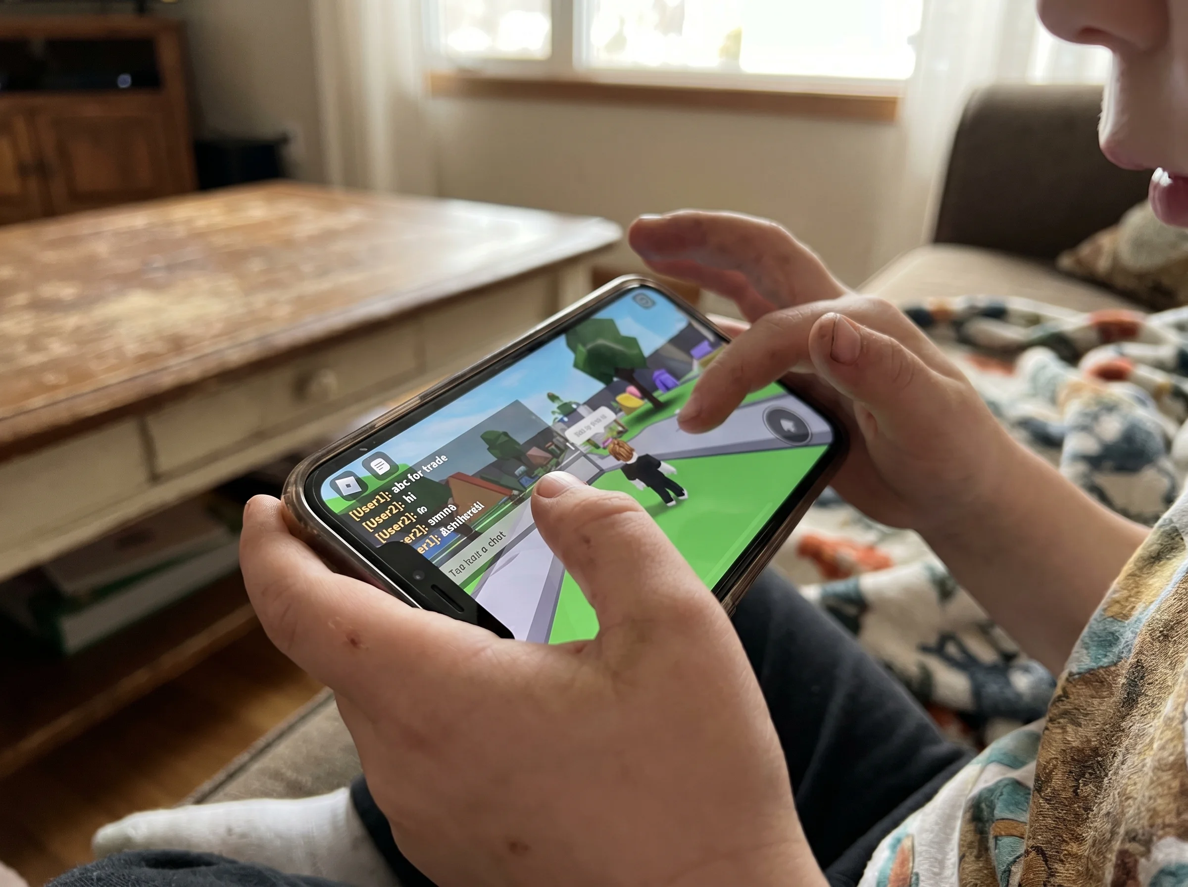 Close-up of a kid's hands holding a phone with a Roblox game on screen, text chat visible in the corner