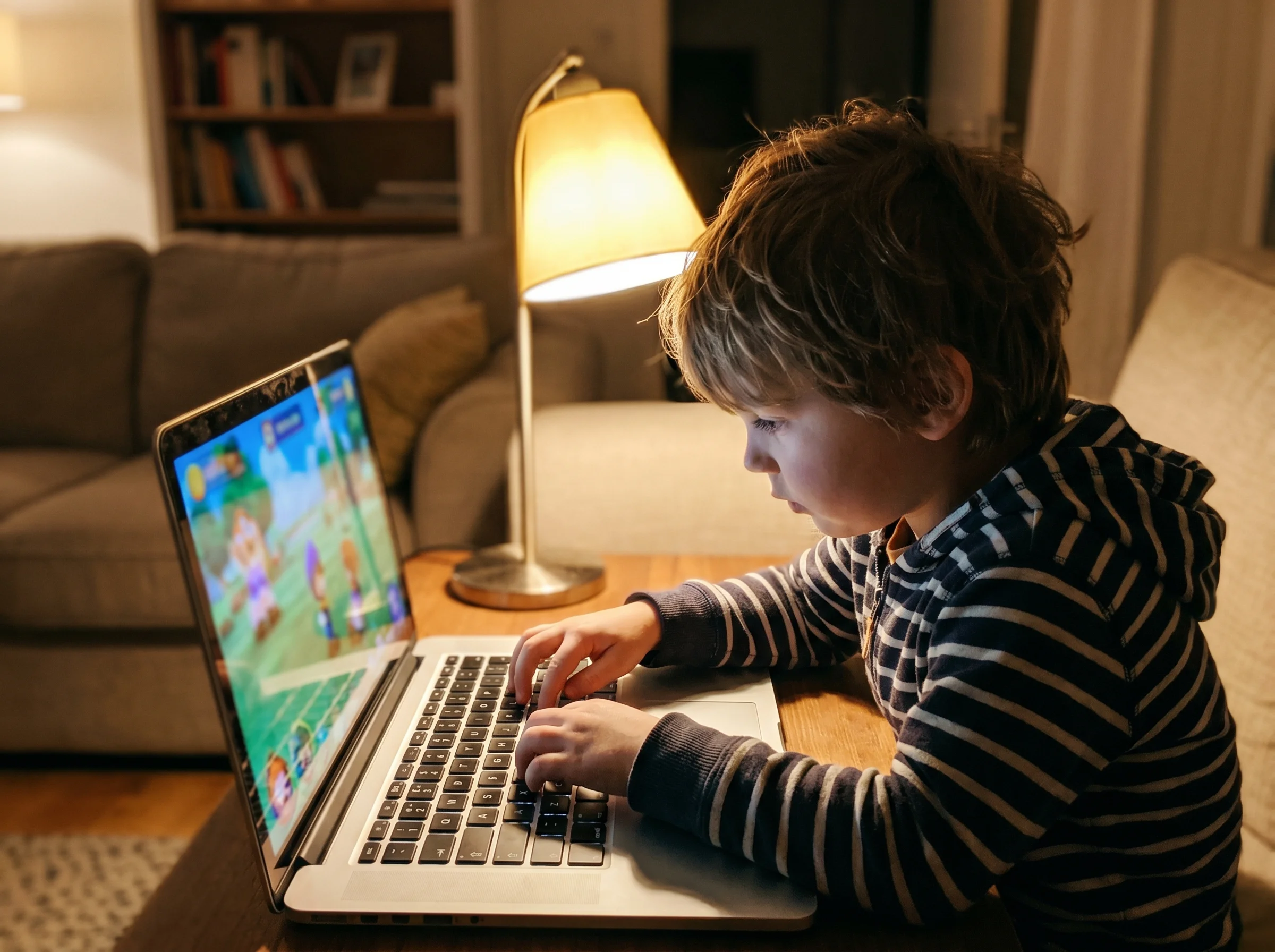 Kid typing on a laptop keyboard, game screen visible but slightly out of focus, evening lamp light