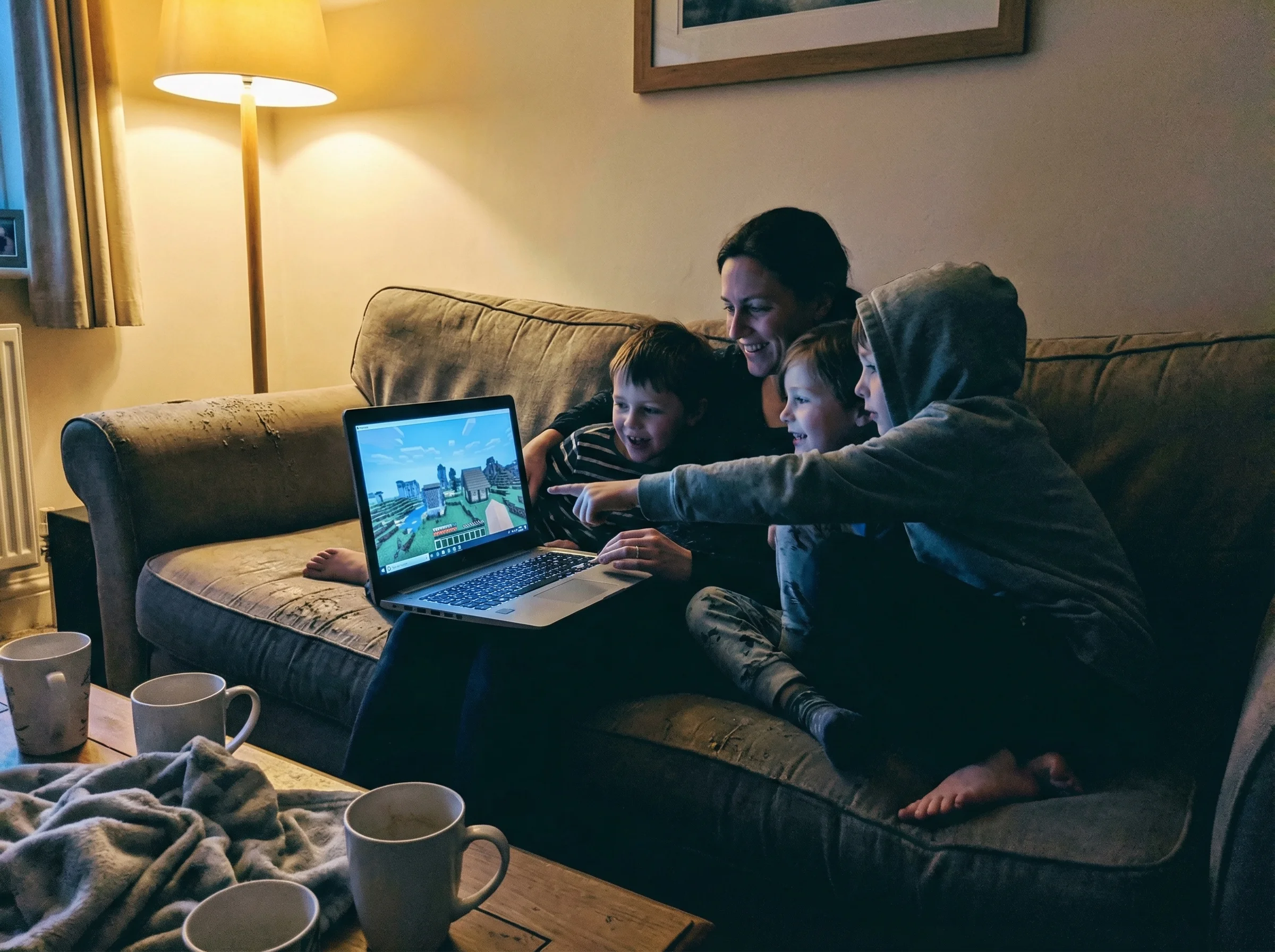 Evening scene with parent and two kids huddled around a laptop, all three faces lit by the screen's glow, showing different M