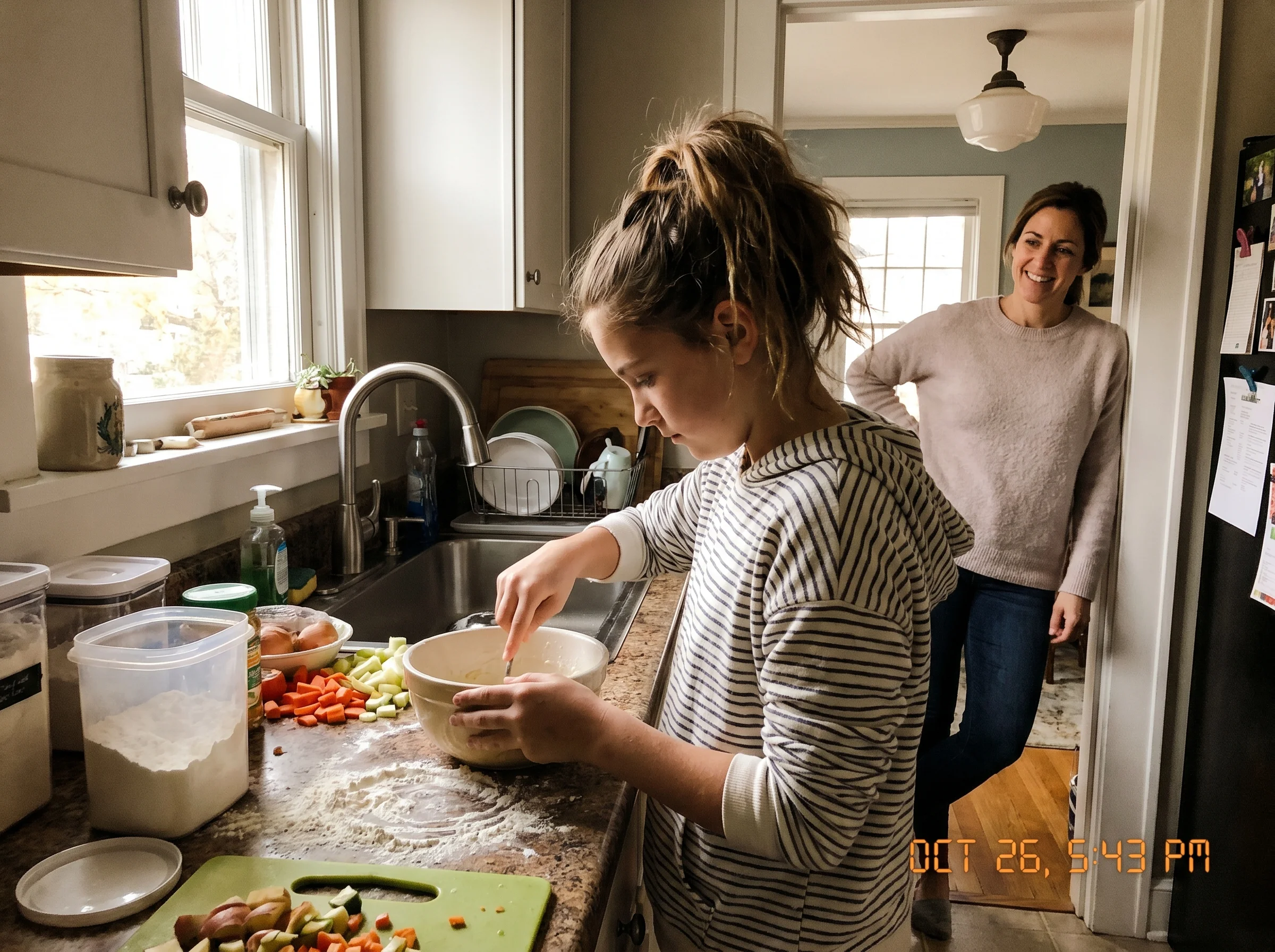 A tween standing at a kitchen counter mid-cook, ingredients spread out, focused expression with a parent watching from the do