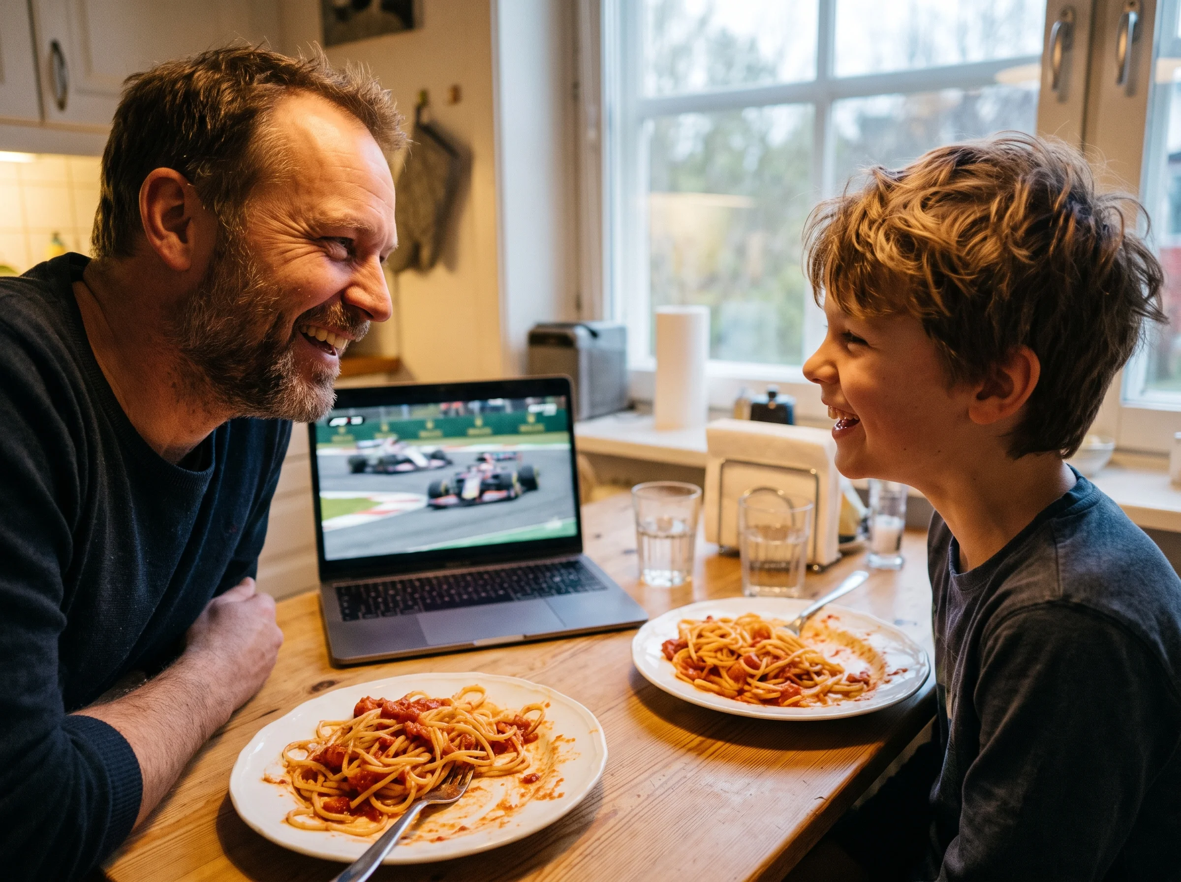 A father and son sharing a laugh at the dinner table with a laptop nearby showing racing highlights, plates of pasta in front