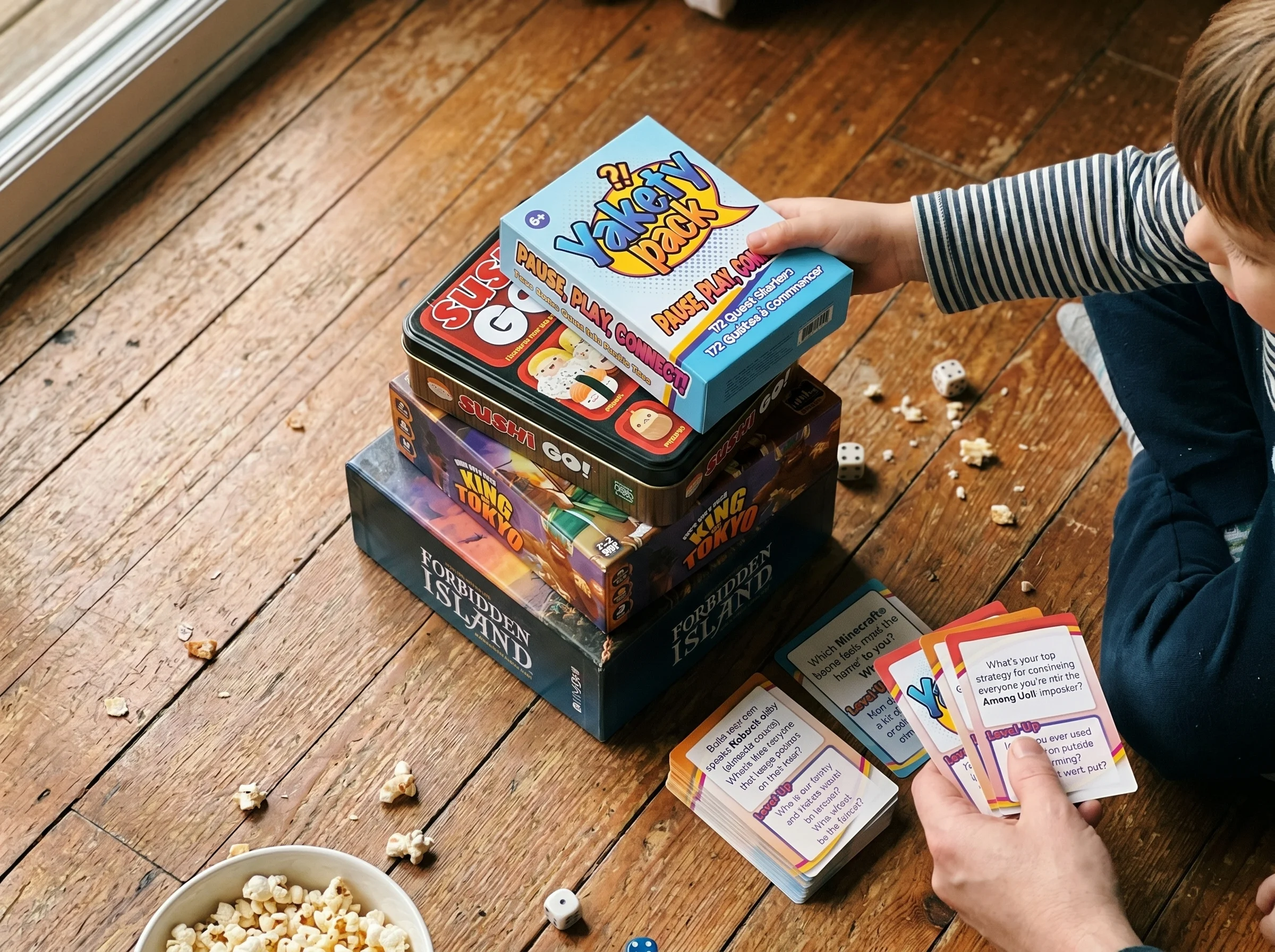 Overhead shot of several board game boxes stacked on a wooden floor, including Sushi Go, King of Tokyo, and Forbidden Island,