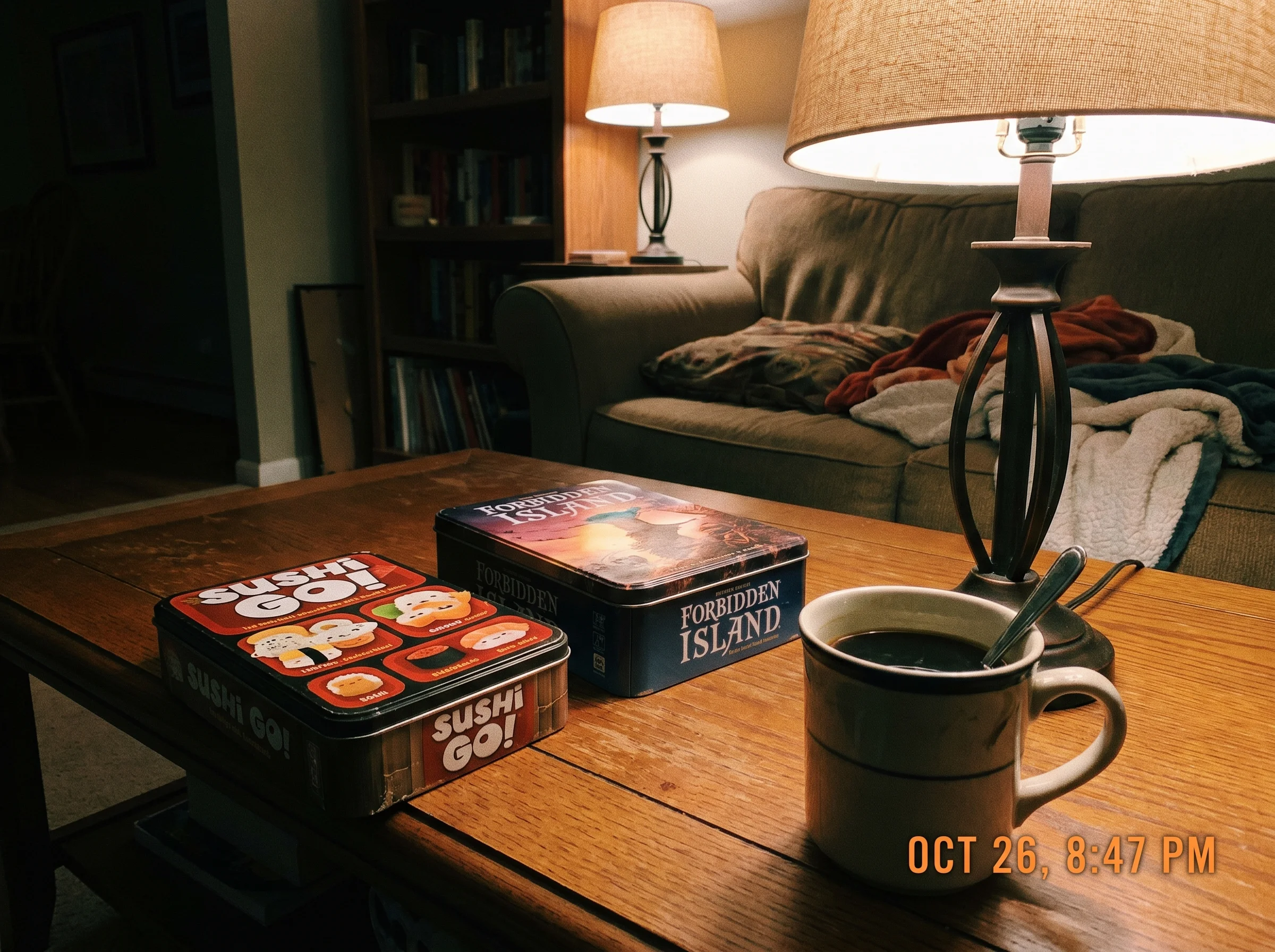 A copy of Sushi Go and Forbidden Island side by side on a coffee table, a mug of coffee next to them, casual evening lamp lig