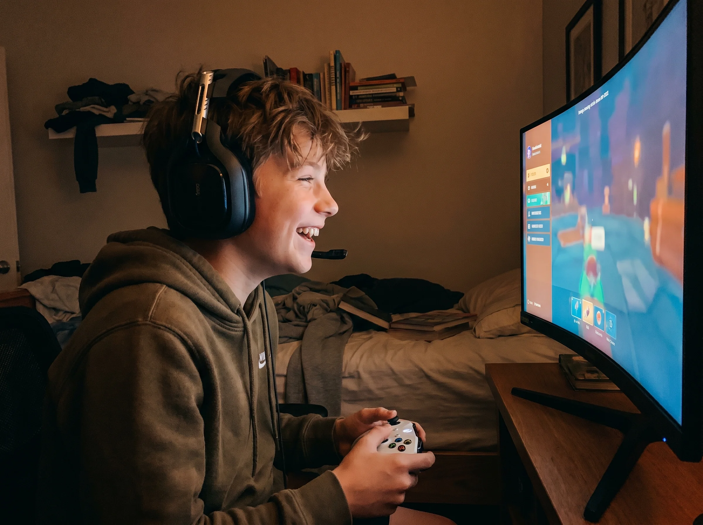 A middle school aged boy with a gaming headset on, laughing at his screen, controller in hand, bedroom lit by the glow of the