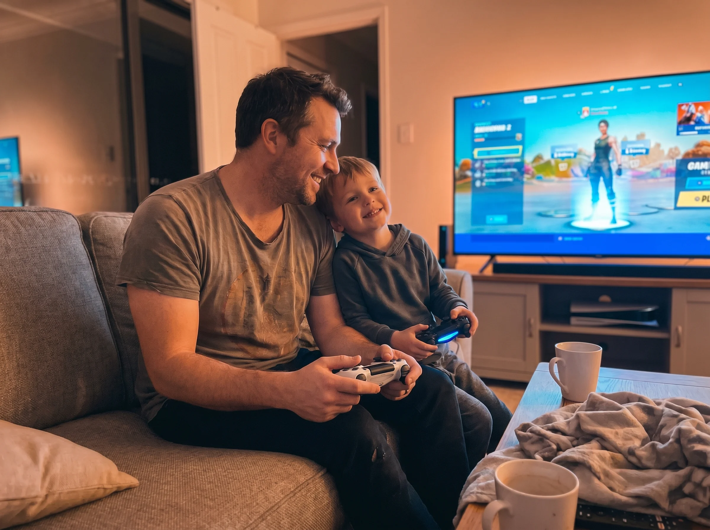 A dad and son sitting on a couch shoulder to shoulder, controllers in hand, screen glow on their faces, casual easy moment, i